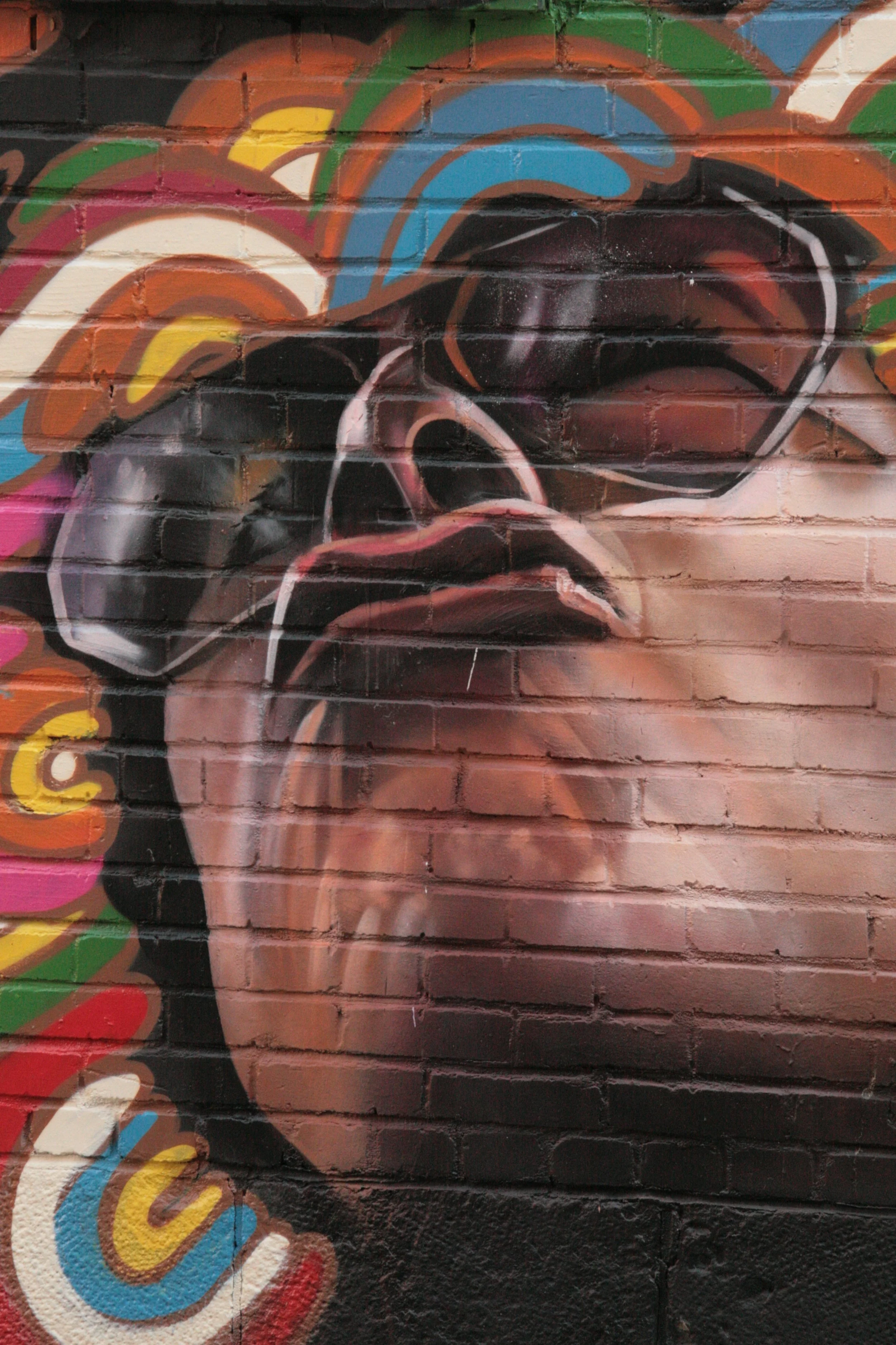 Graffiti mural of a woman's face wearing sunglasses, with colorful swirling patterns around her head painted on a brick wall.
