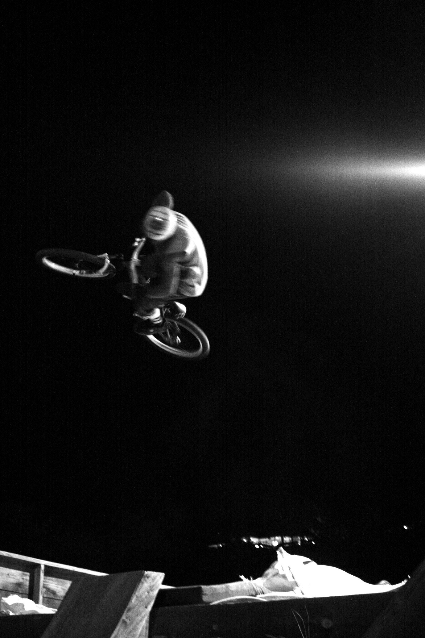 Black-and-white photo of a person performing a BMX stunt in mid-air at night, with a beam of light shining in the background.