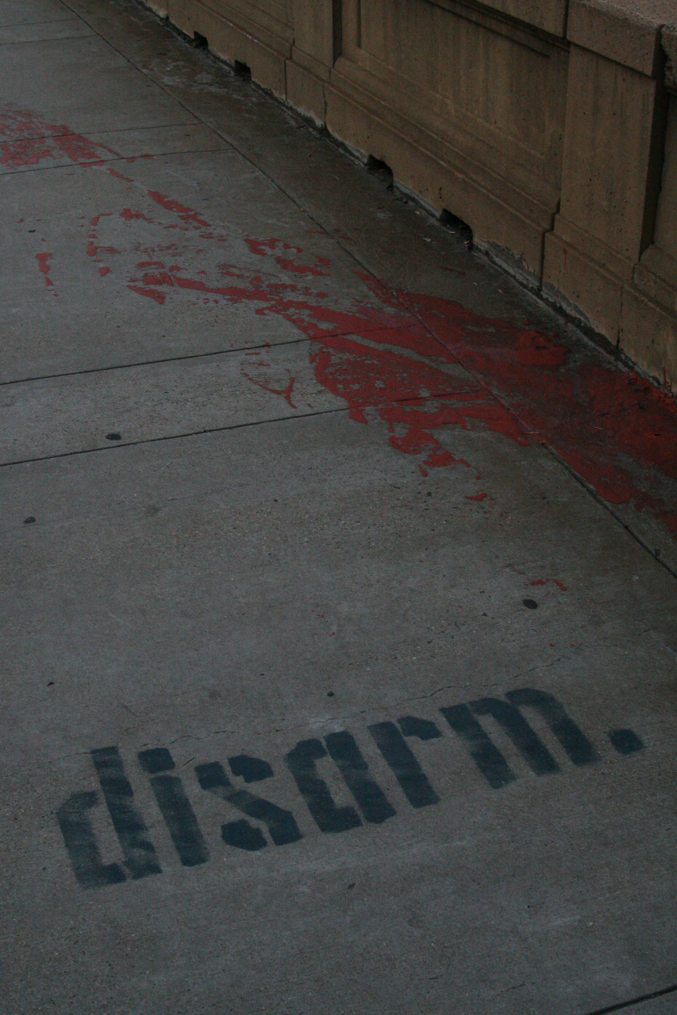 A sidewalk with red paint splattered near the wall and a stenciled warning sign that says 'CAUTION' in black letters.