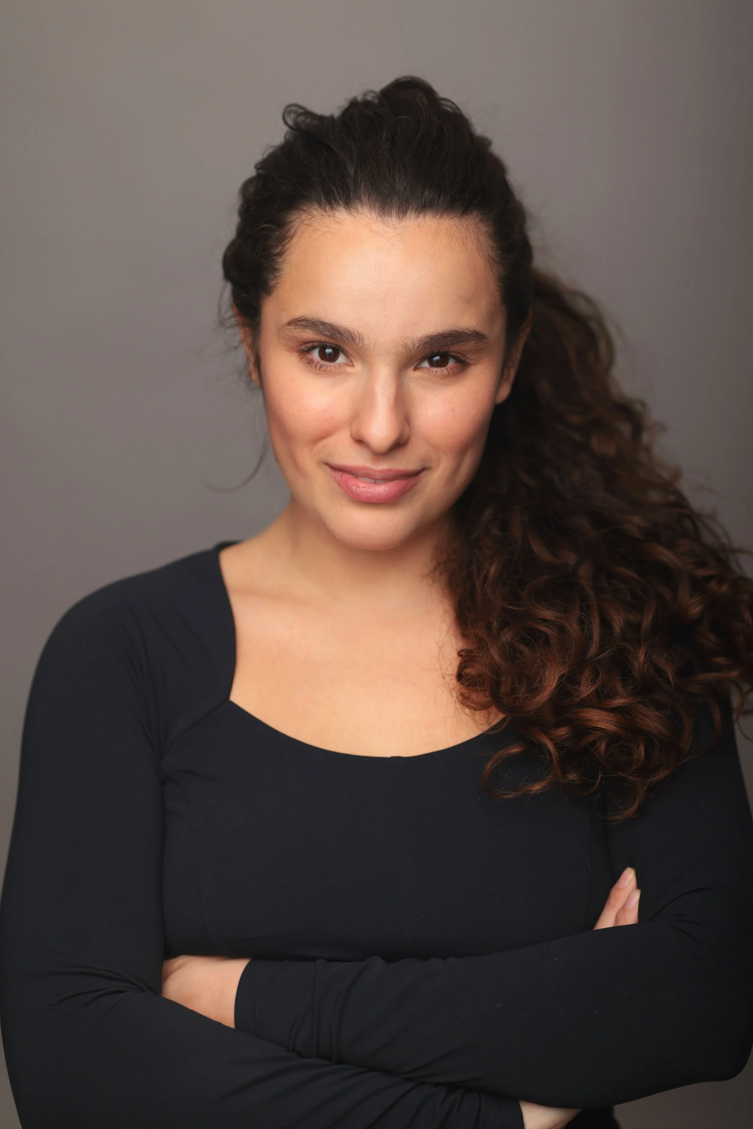 Portrait of a young woman with long, curly brown hair, wearing a black top, smiling softly against a gray background.