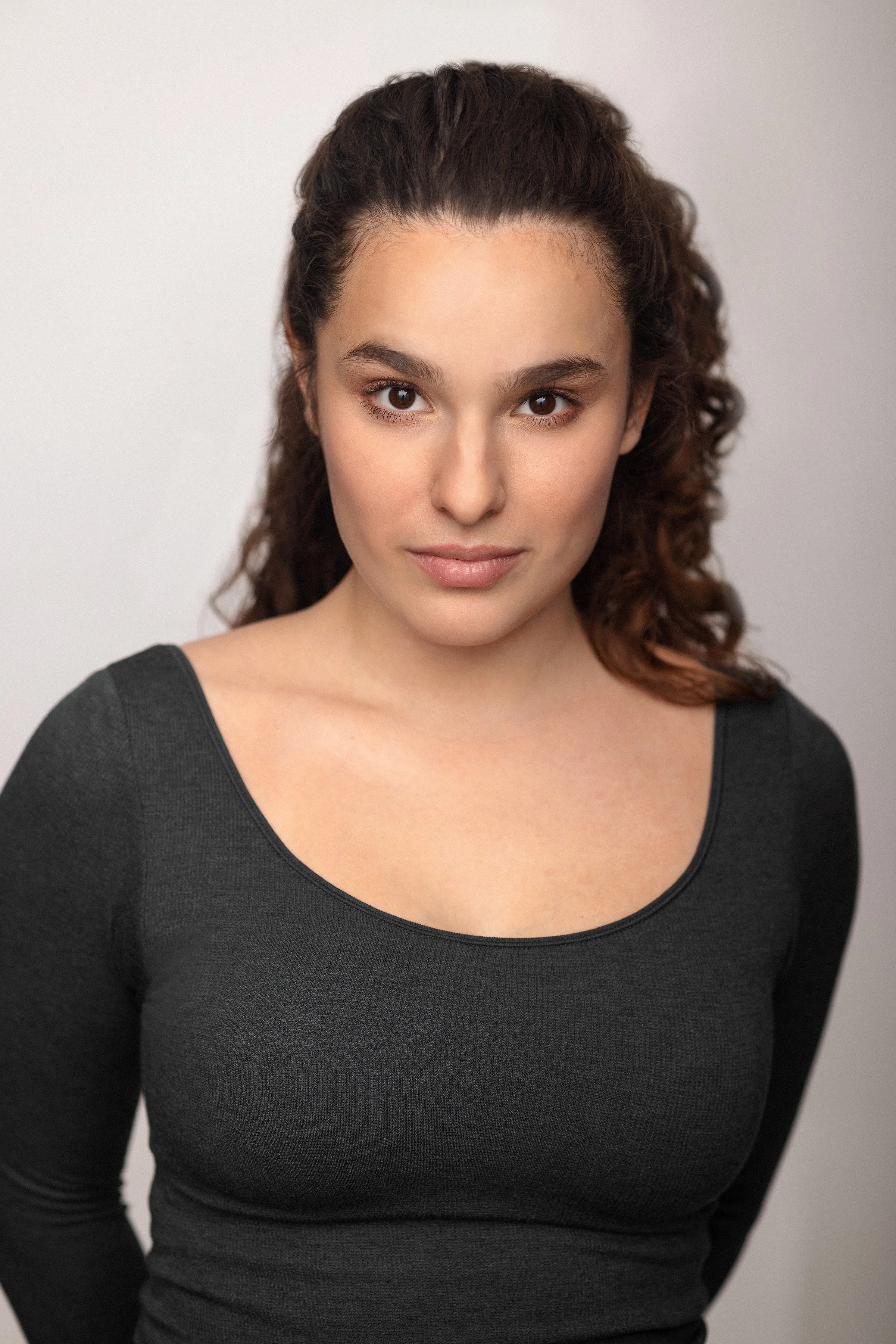 A young woman with curly brown hair, wearing a black long-sleeve top, looking directly at the camera with a neutral expression.