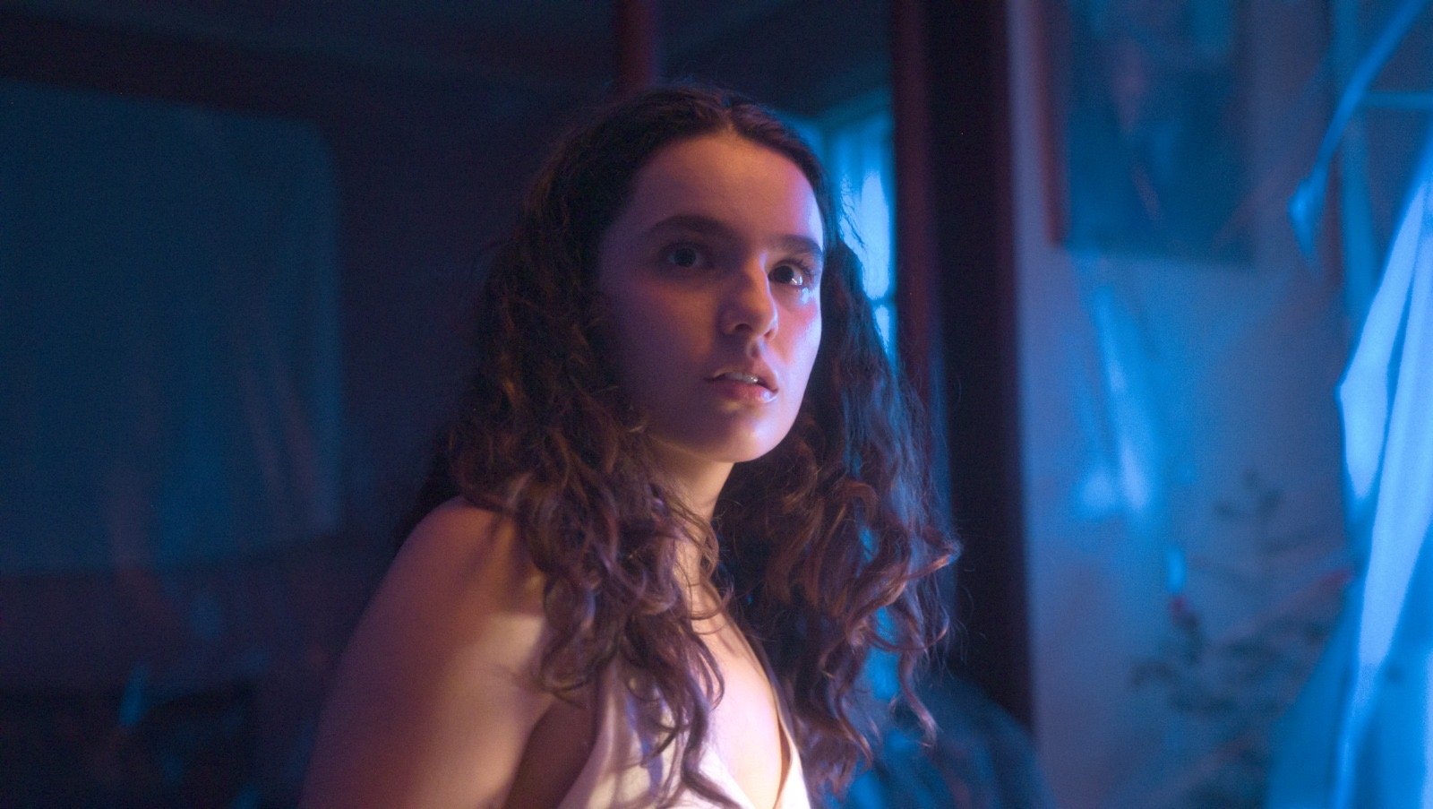 A young woman with curly brown hair and fair skin, looking over her shoulder, in a dimly lit room with blue and purple hues.