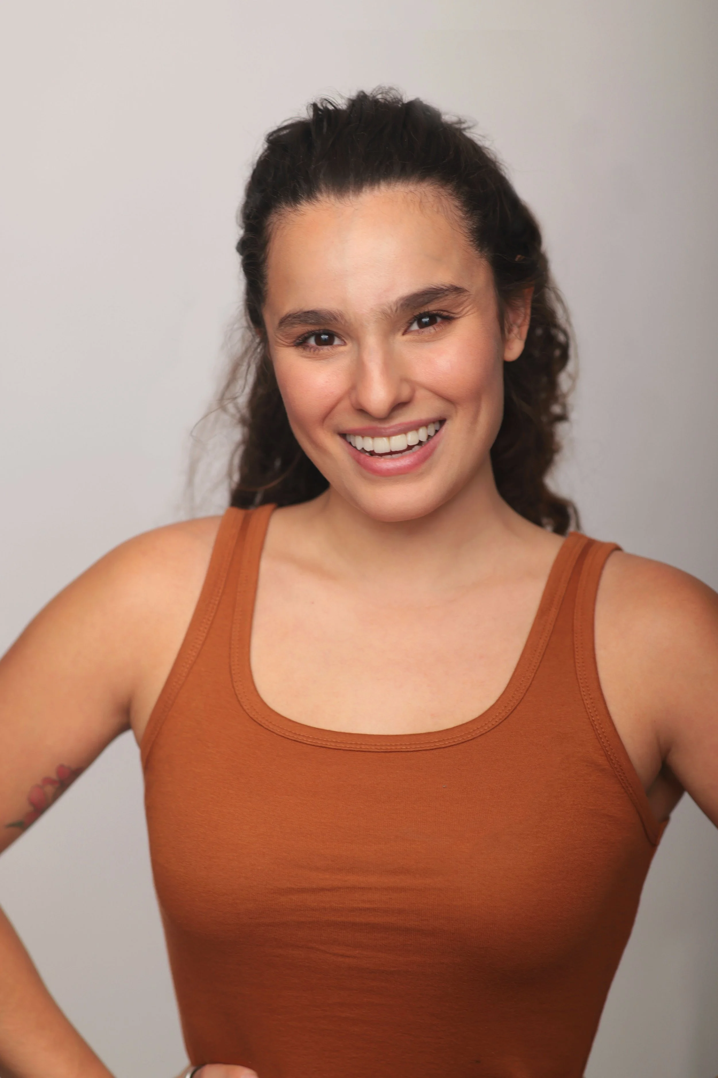A woman with long, curly brown hair smiling at the camera, wearing an orange tank top against a plain background.