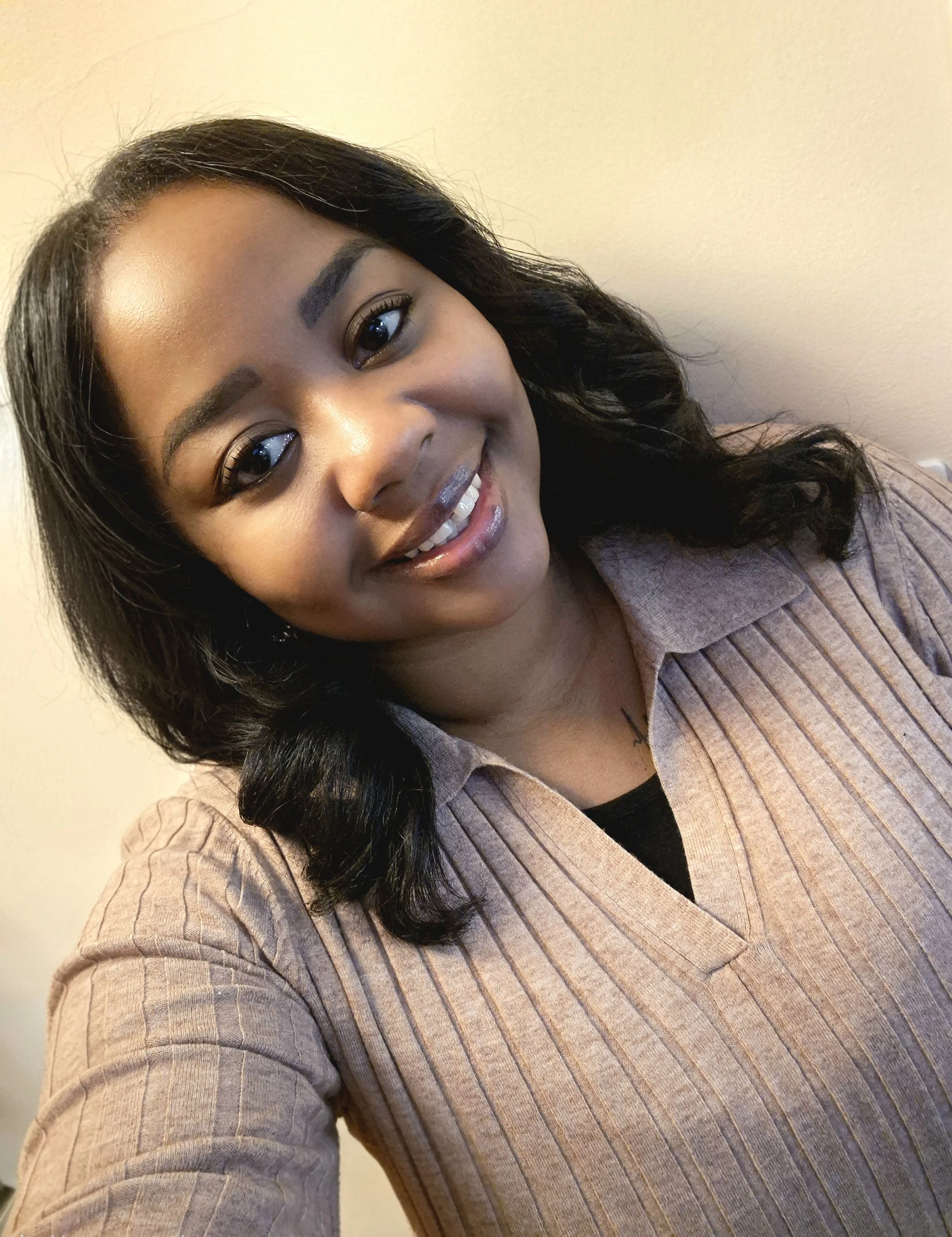 A young woman with dark hair and a friendly smile, wearing a beige ribbed sweater over a black top, standing against a plain wall.