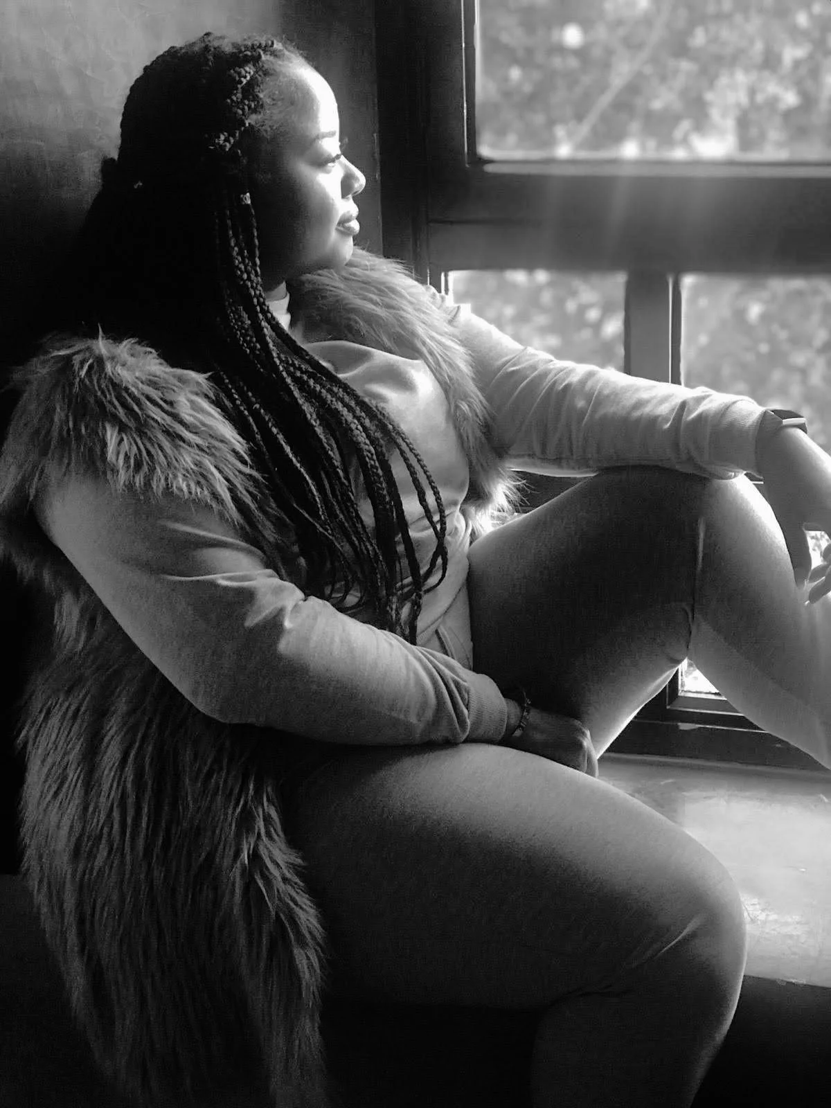A woman with braided hair sitting by a window, gazing outside, with a fur coat over her shoulders.