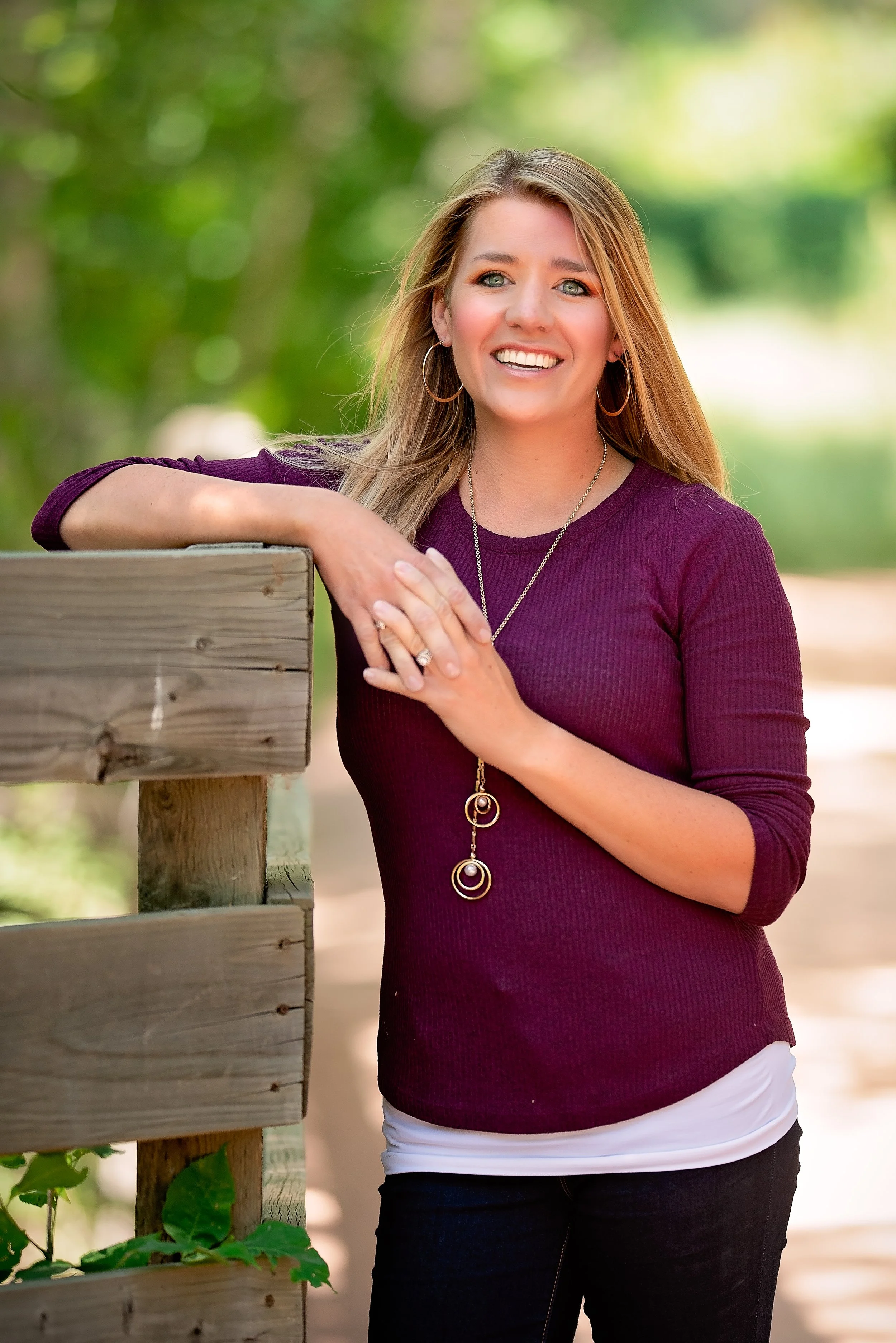A woman with blonde hair and hoop earrings smiling outdoors, leaning on a wooden railing, wearing a maroon long-sleeve shirt and a necklace with circular pendants.