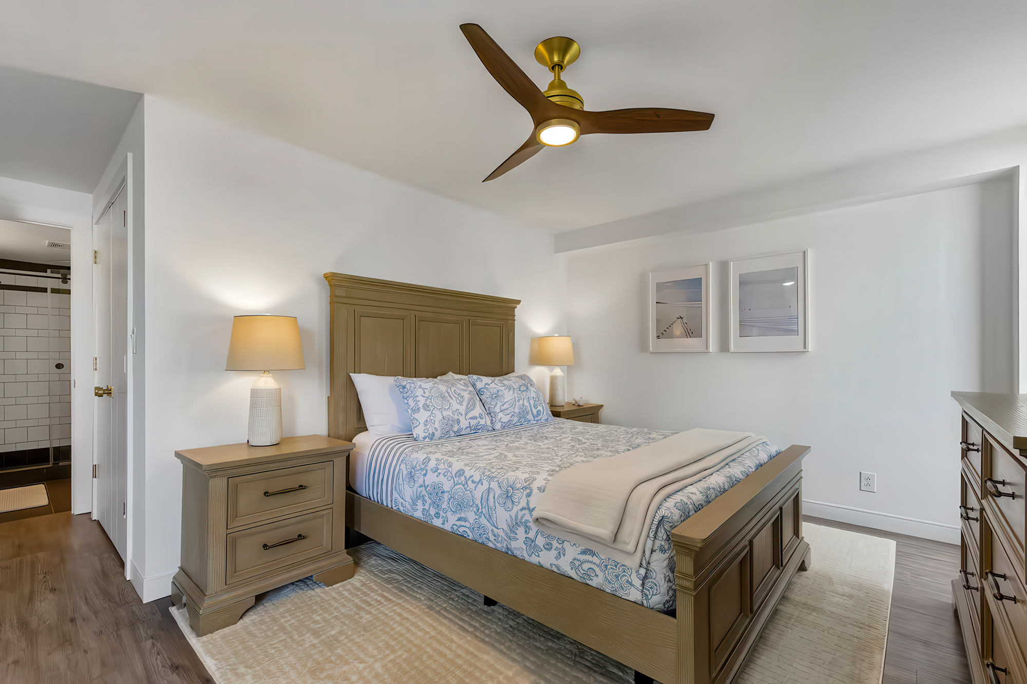 A bedroom with a beige bed frame, white and blue patterned bedding, two matching bedside nightstands with table lamps, a wood ceiling fan with LED light, and framed artwork on the white wall. Part of an adjacent bathroom with a tile shower is visible through the door.