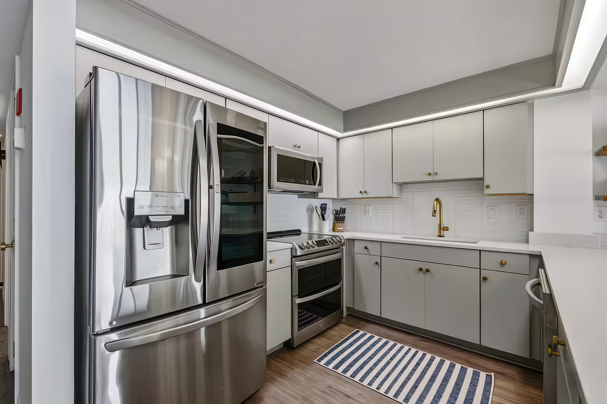 Modern kitchen with white cabinets, stainless steel refrigerator, microwave, oven, and small area rug on hardwood floor.