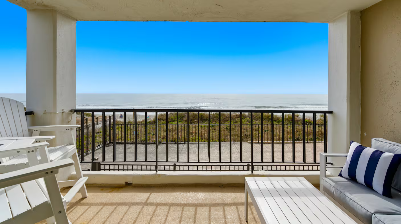 Beach view from a balcony with white chairs and a small table, black railing, and ocean in the distance under a blue sky.