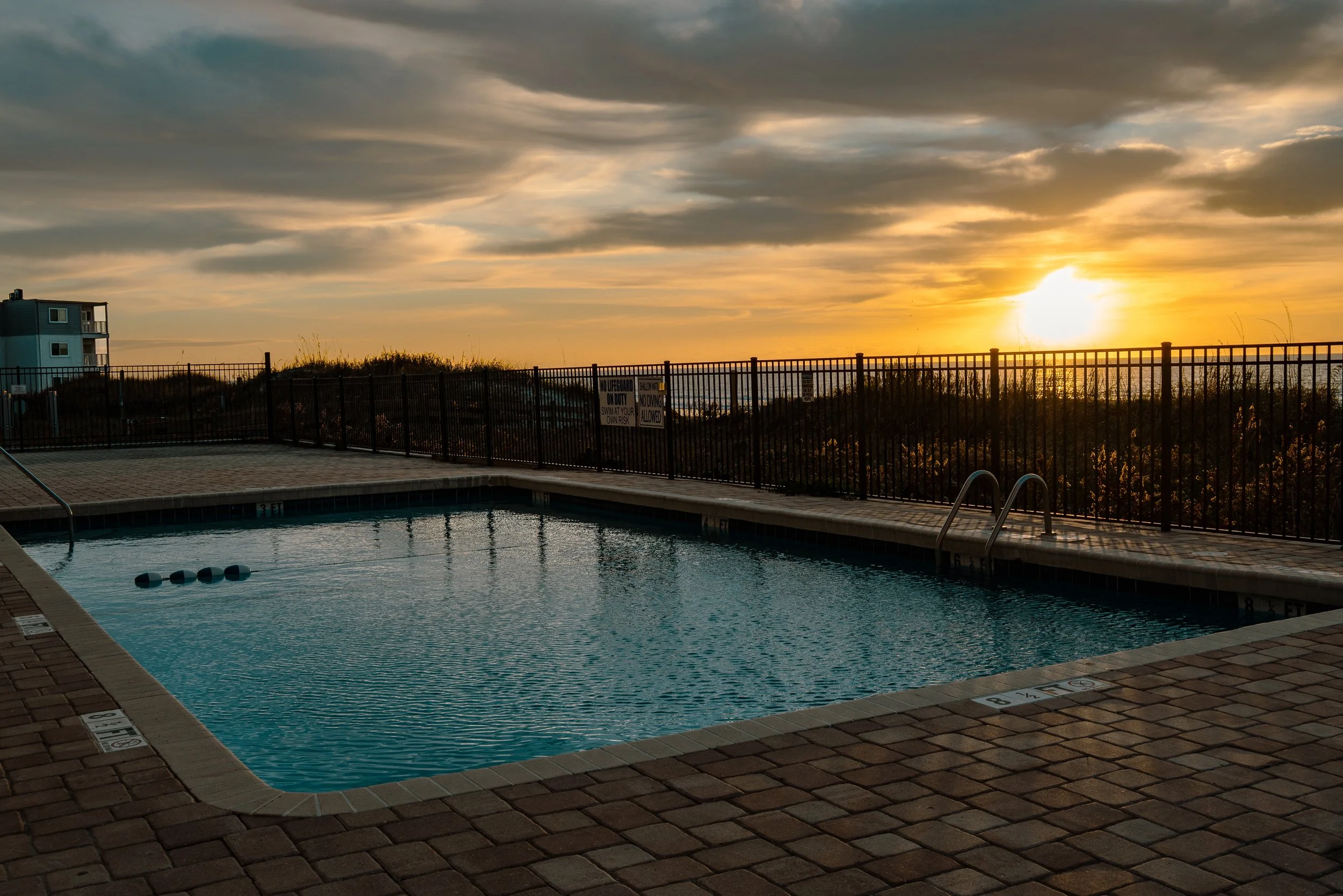 An outdoor swimming pool at sunset with a black metal fence, and a cloudy sky with the sun setting in the background.