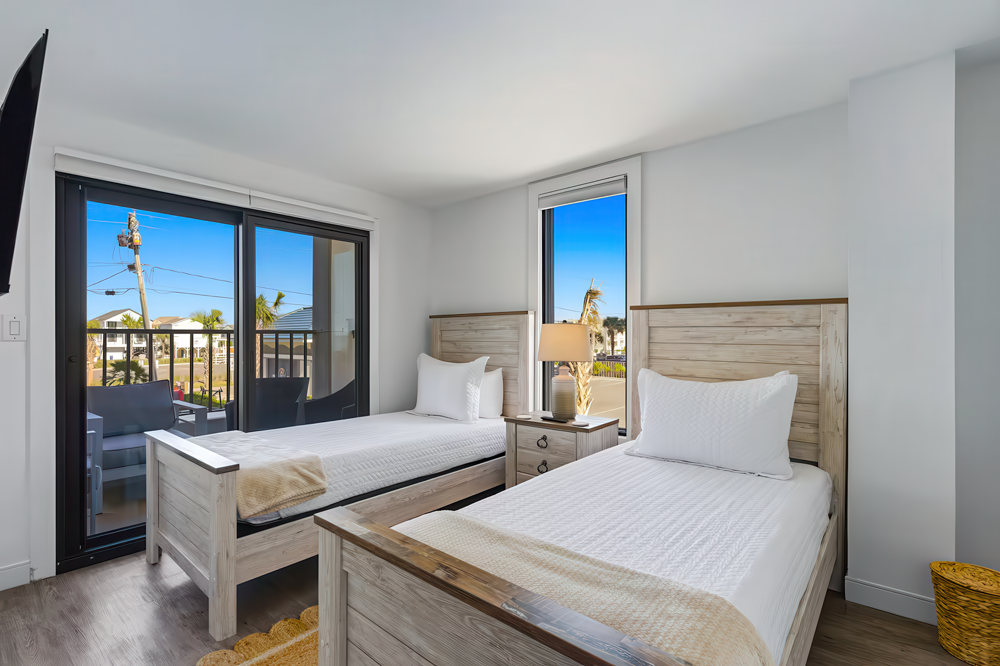 A bright hotel room with two twin beds and light wood headboards. Large windows and a sliding door to a balcony show a sunny exterior with palm trees and a blue sky. The room is decorated in white with minimalistic furniture, including a lamp on a small nightstand and a yellow basket on the floor.
