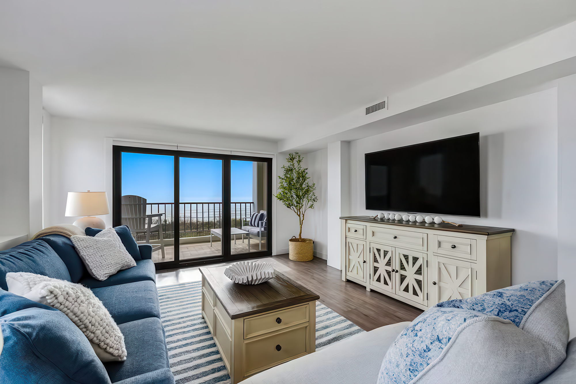 Bright living room with blue sofas, decorative pillows, a wooden coffee table, a white TV console, a large flat-screen TV, a potted plant, a striped rug, and sliding glass doors leading to a balcony with outdoor furniture and a scenic view.