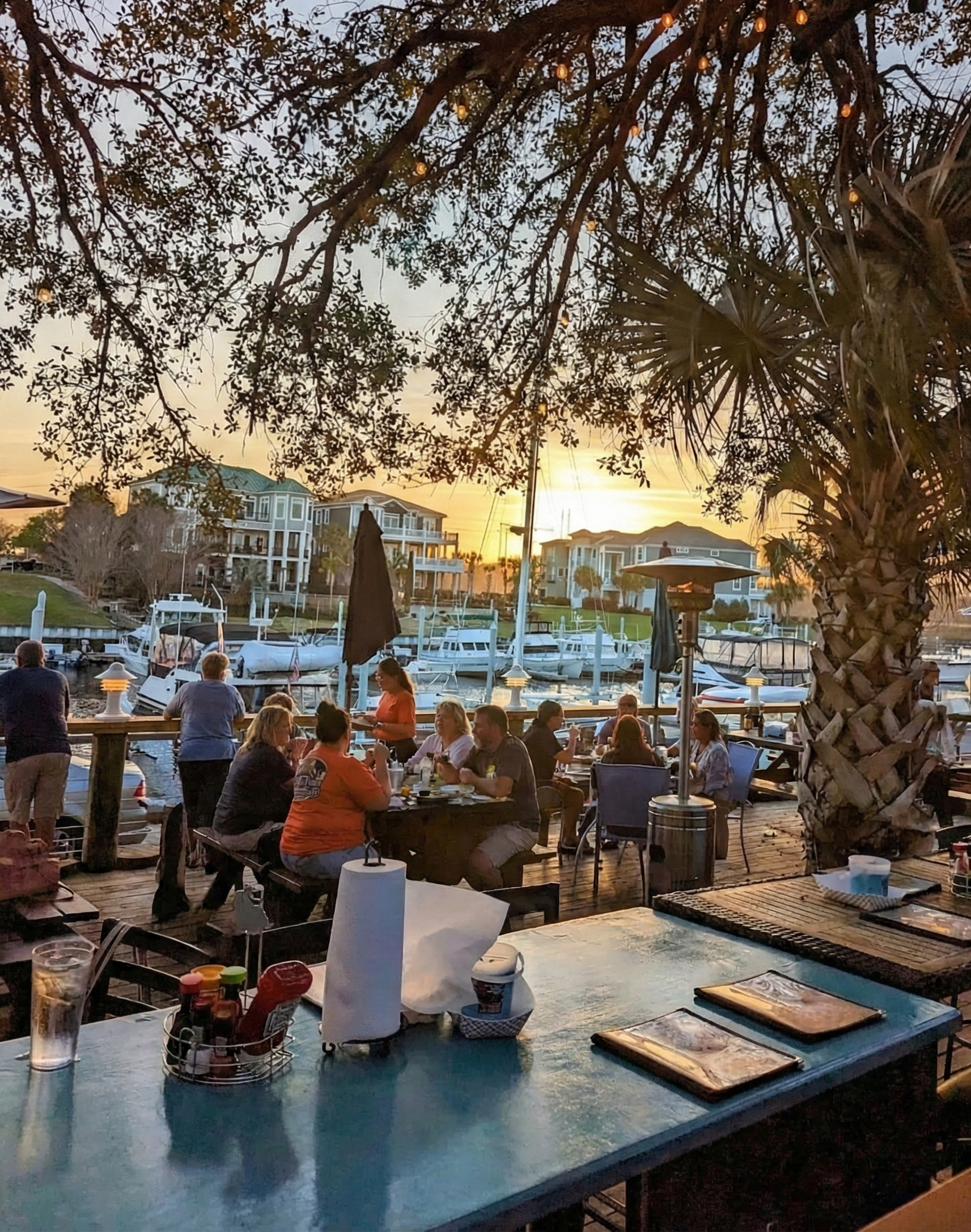 People dining at an outdoor restaurant by a marina during sunset, with boats docked and residential buildings in the background, under the shade of a large palm tree.