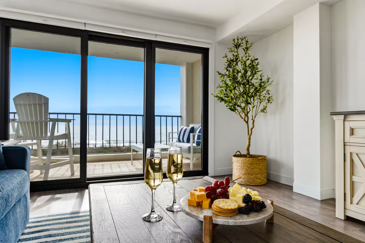 A living room with sliding glass doors opening to a balcony view of the ocean. The room has a wooden table with two glasses of champagne, a cheese platter, and a striped rug. There is a potted tree in the corner and a white cabinet on the right.