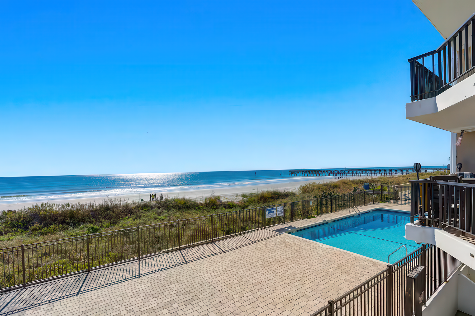 View of a beachfront property with a swimming pool, outdoor patio, and a white building with balconies overlooking the ocean and a pier extending into the water under a clear blue sky.