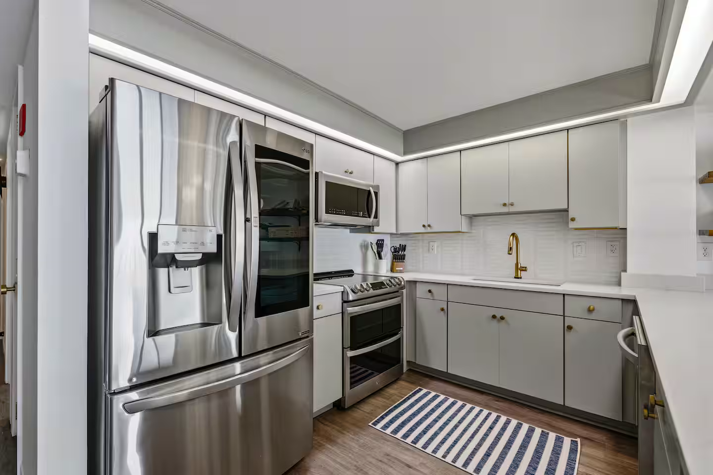 Kitchen with stainless steel LG refrigerator, microwave, stove, and white cabinets with gold knobs, gold faucet, and a striped rug on wooden floor.