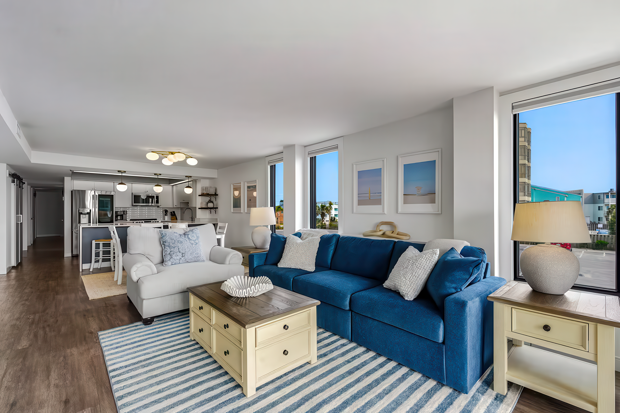Open-concept living room with blue sofa, white armchair, wooden coffee table, area rug, and large windows showing a cityscape.