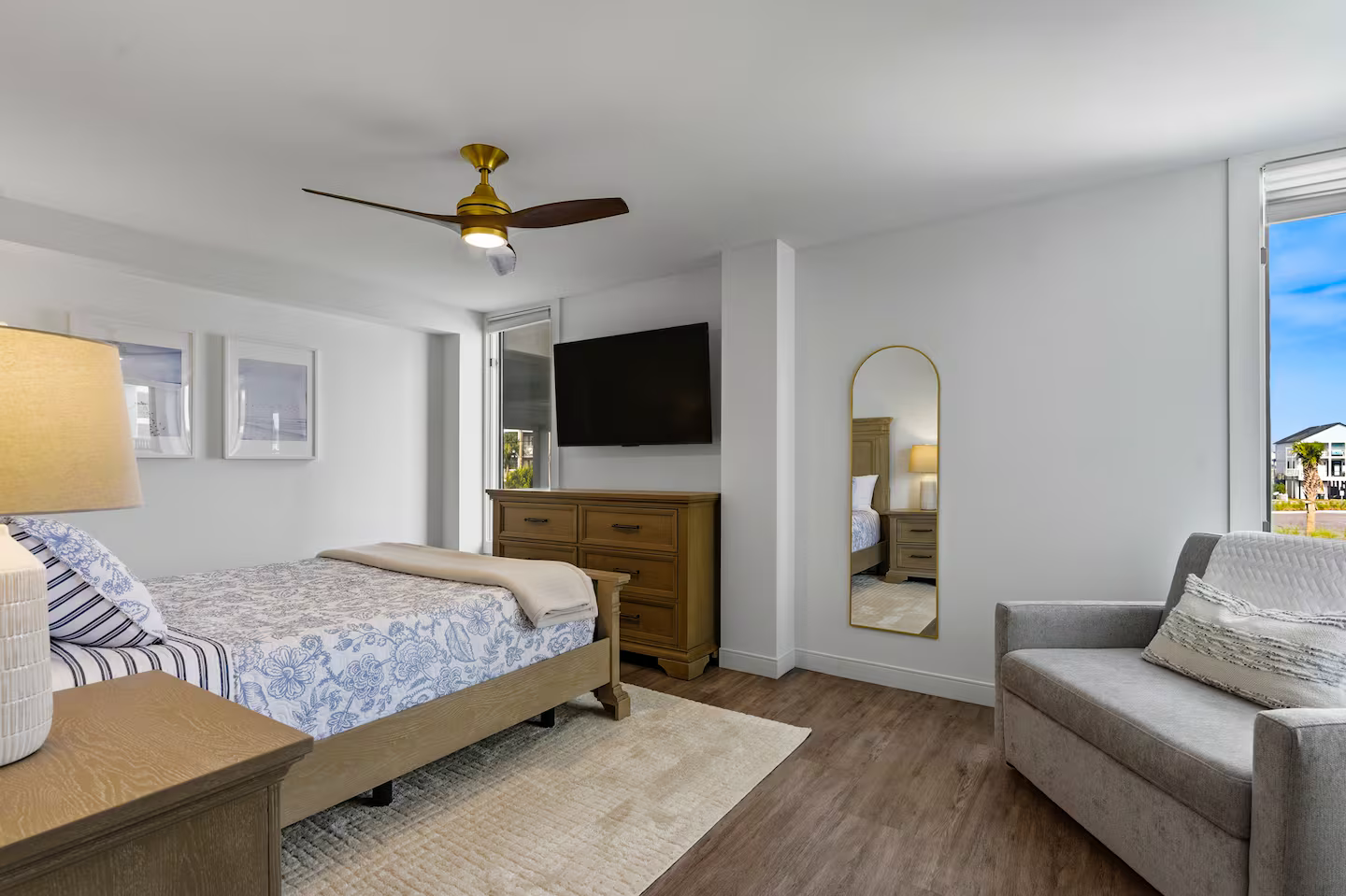 Modern bedroom with white walls, wooden furniture, a bed with blue and white linens, a gray armchair, a flat-screen TV, a mirror, a ceiling fan, and large windows showing an outdoor scene.