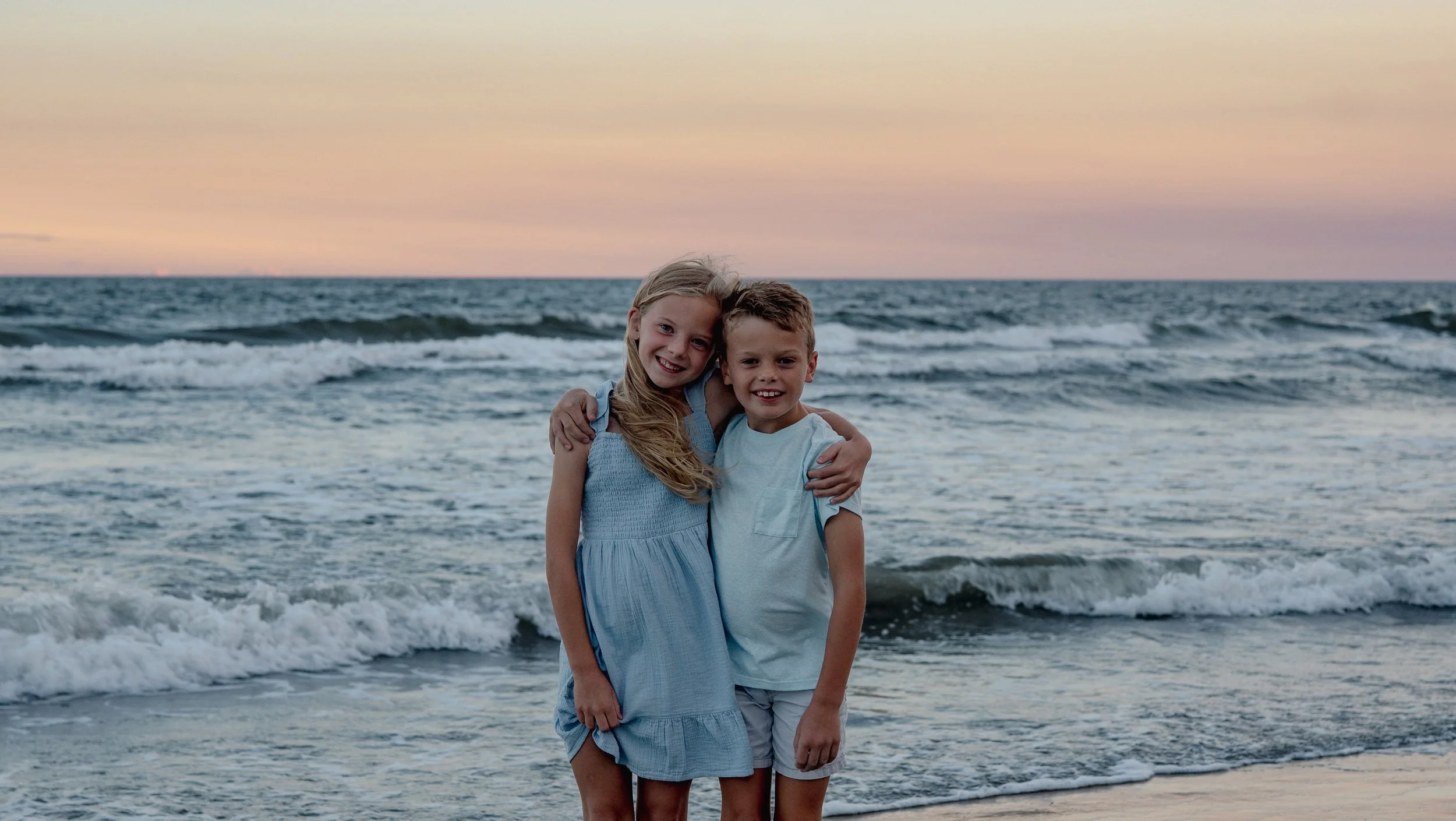 A young girl and boy hugging at the beach during sunset with ocean waves in the background.