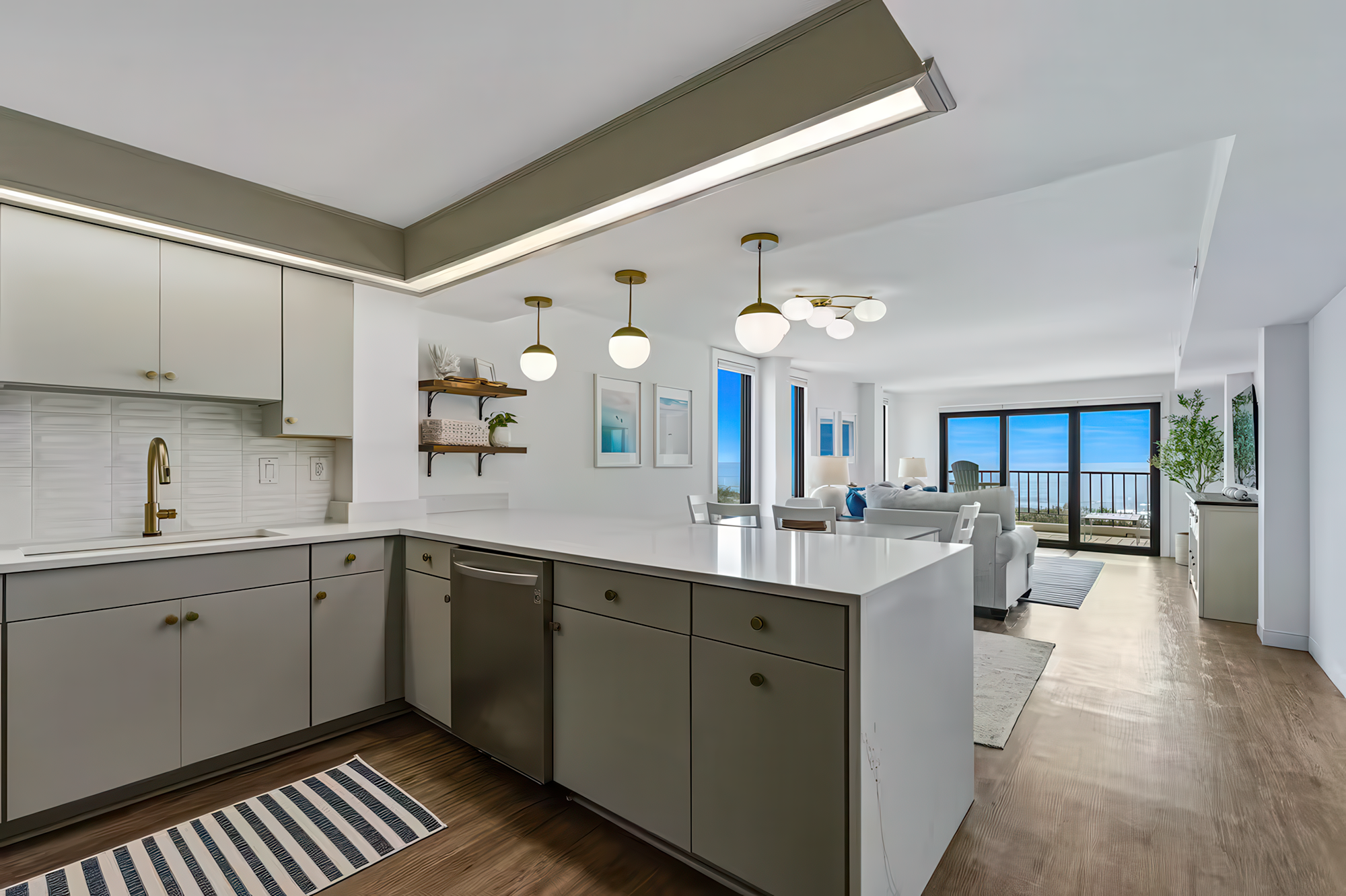 Modern open-concept living space with kitchen in foreground, white cabinetry, gold hardware, and a white quartz island, leading to a living room with white sofa, artwork, and large glass doors with ocean view.