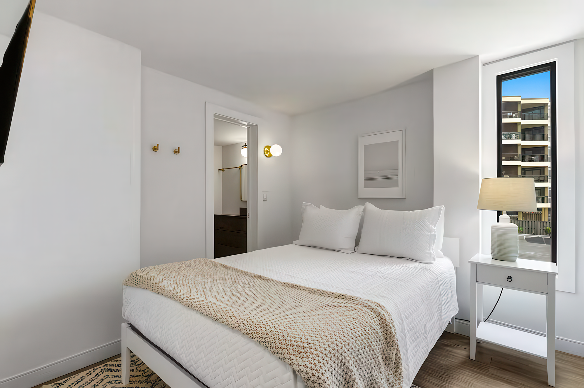Modern bedroom with white walls and bedding, a bedside table with a lamp, a window showing an apartment building outside, and a doorway leading to a bathroom.