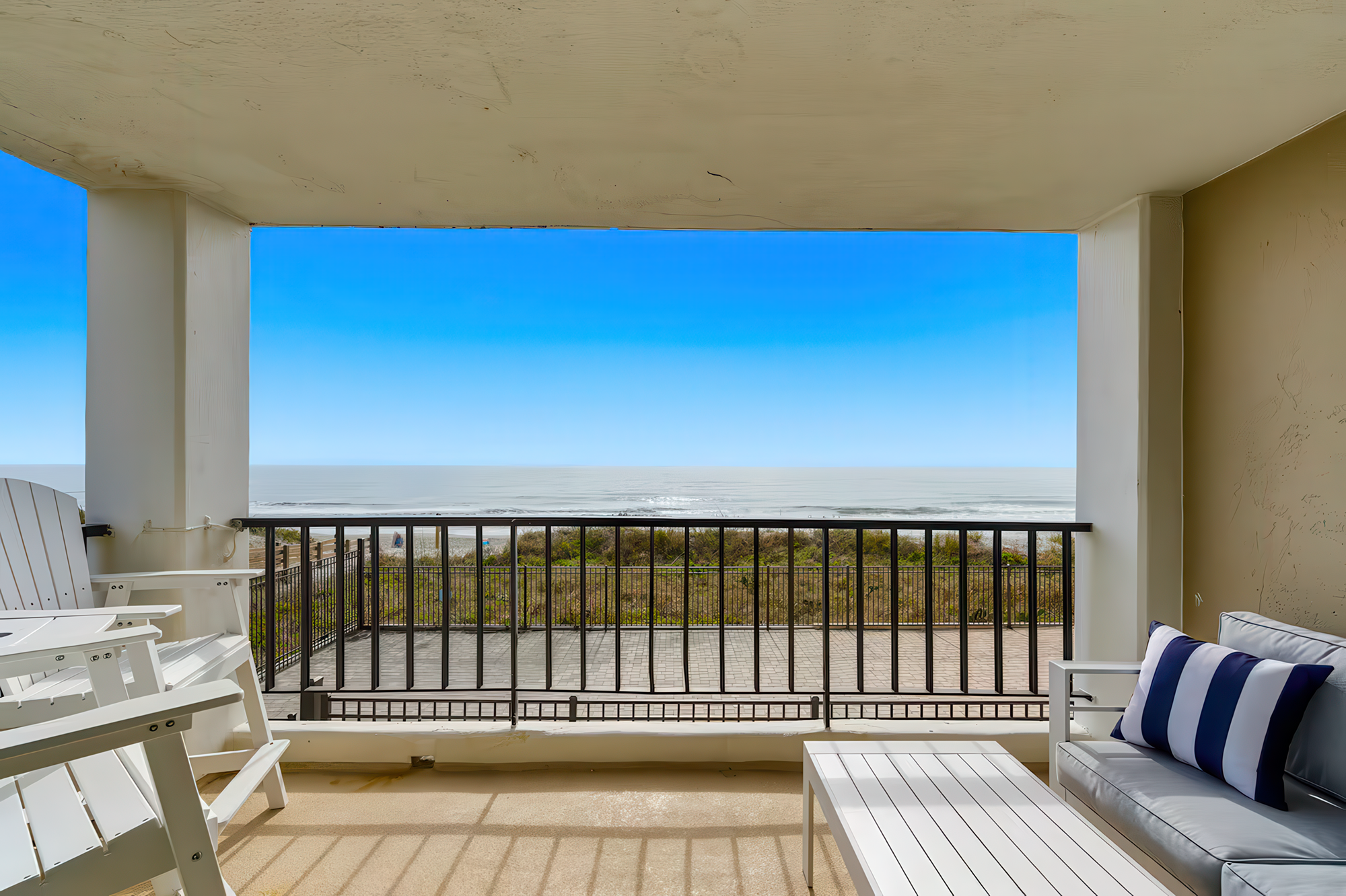 A balcony with white Adirondack chairs, a table, and a cushioned outdoor sofa with navy and white striped pillows, overlooking a beach and the ocean under a clear blue sky.