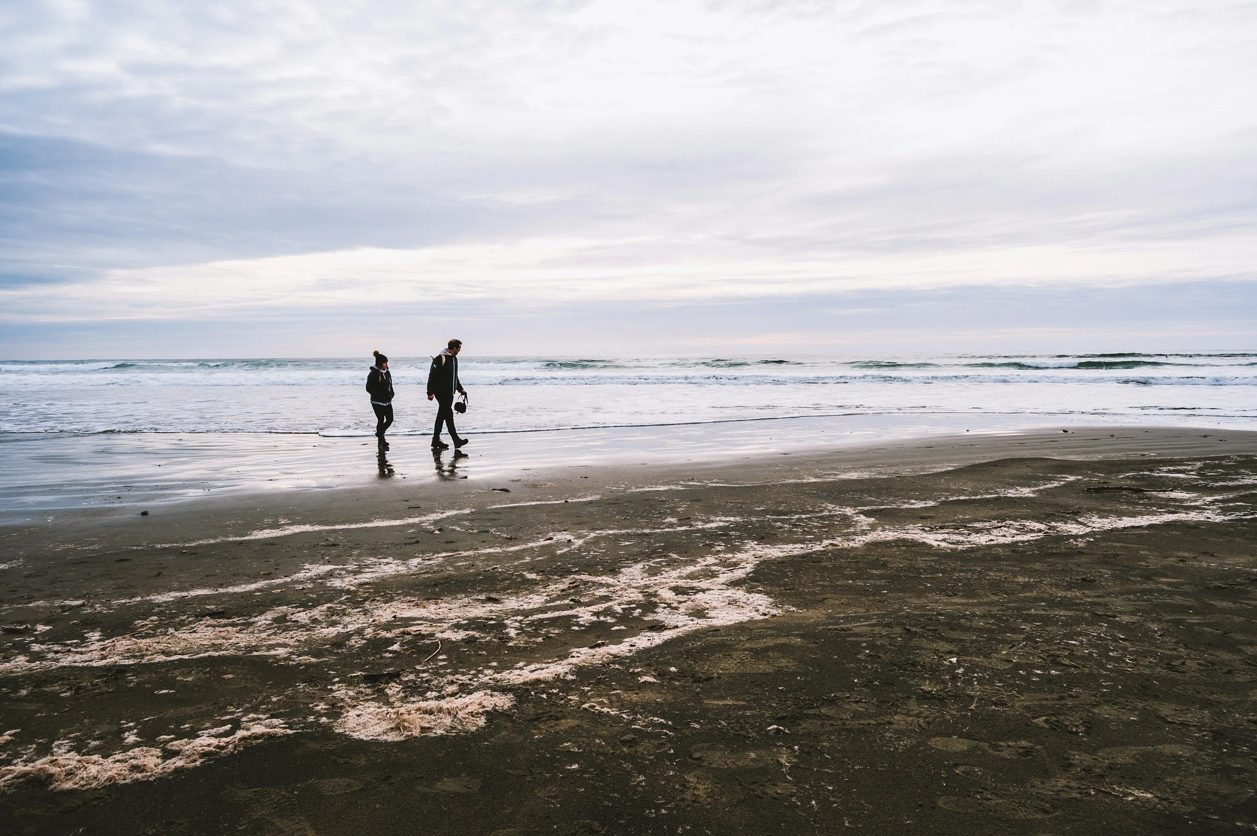 Two people walking along a wet sandy beach near the ocean under a cloudy sky.
