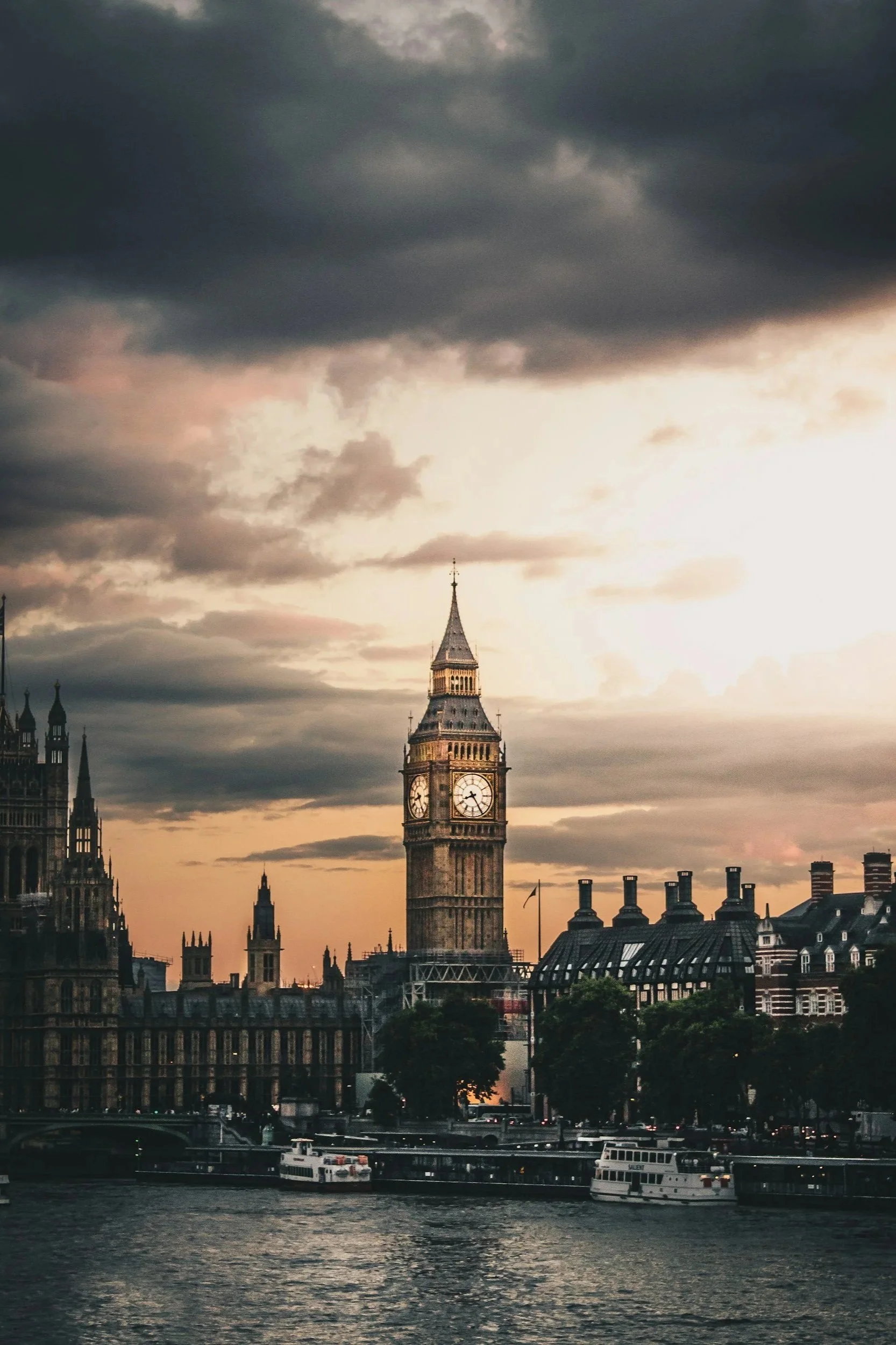 A cityscape featuring the Big Ben clock tower in London during sunset, with cloudy sky, historic buildings, boats on the river, and trees in the foreground.