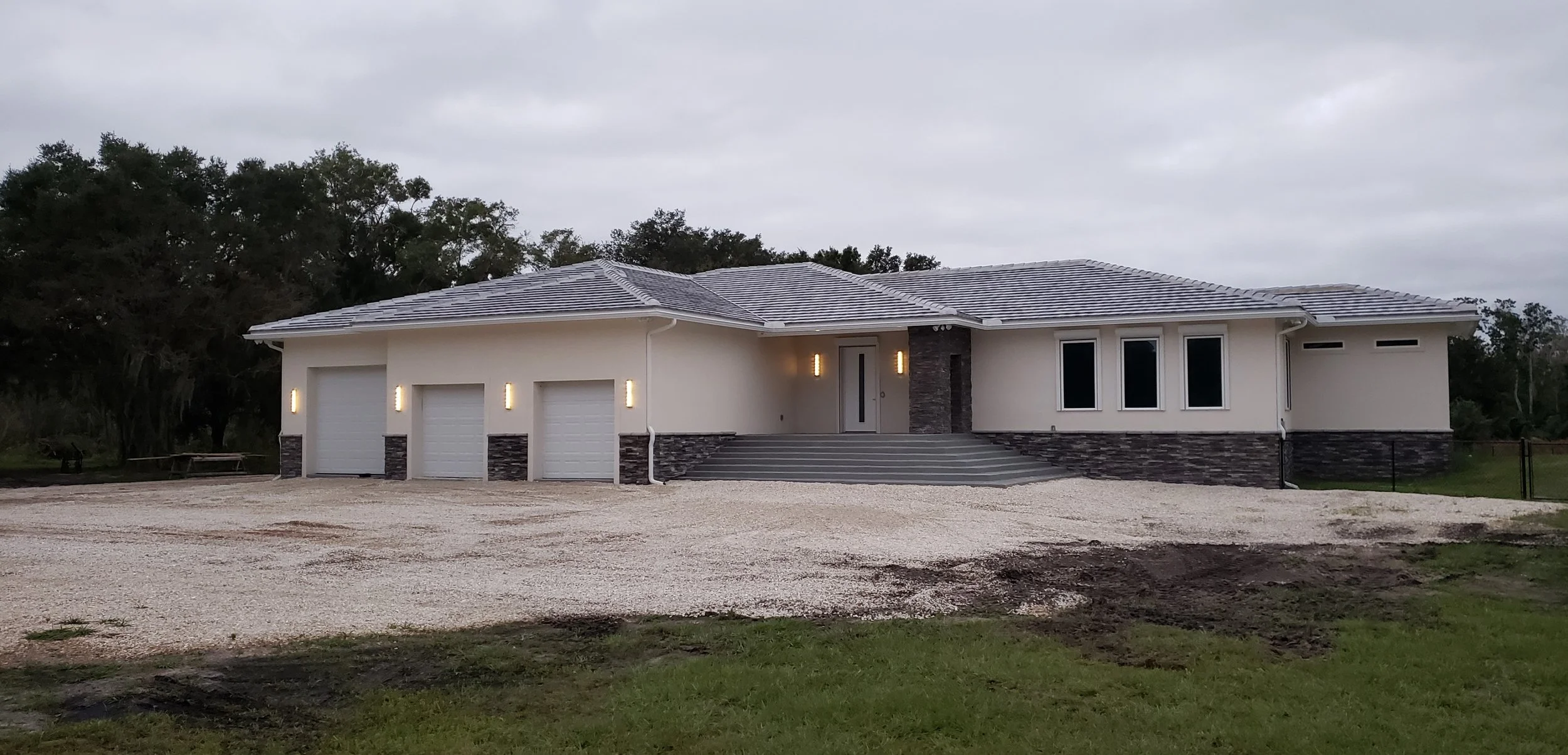 Modern single-story house with three-car garage, stone accents, and a landscaped front entrance with steps, surrounded by a gravel driveway and open space, under a cloudy sky.
