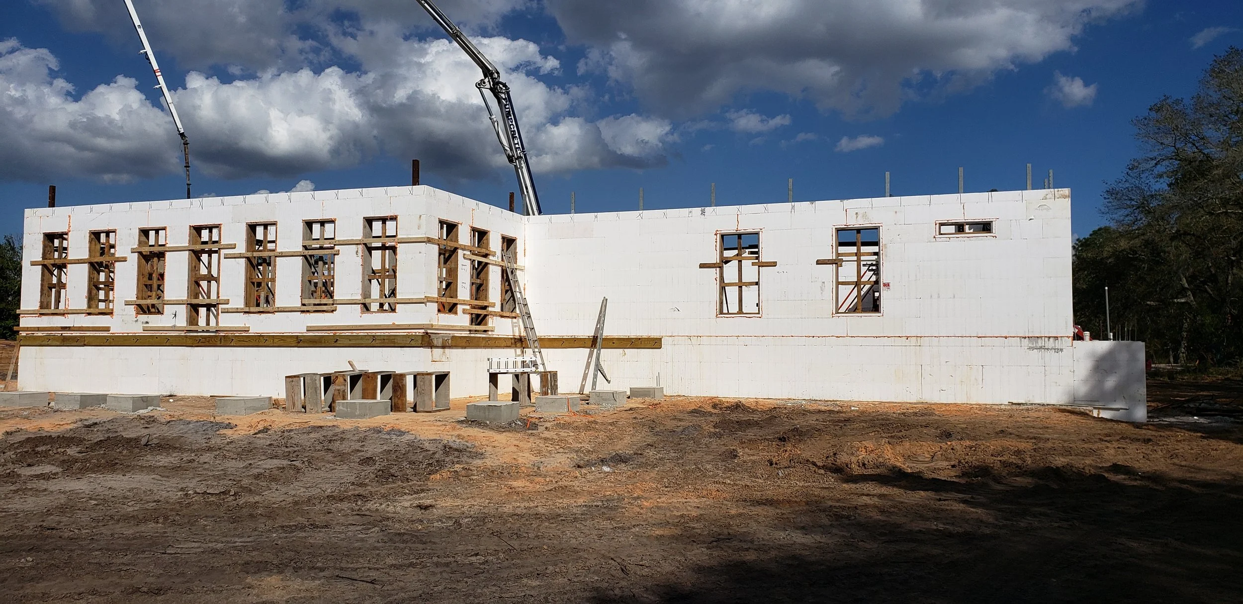 Construction site with a partially built white wall, wooden framing for windows, and a crane arm overhead, under a partly cloudy sky.