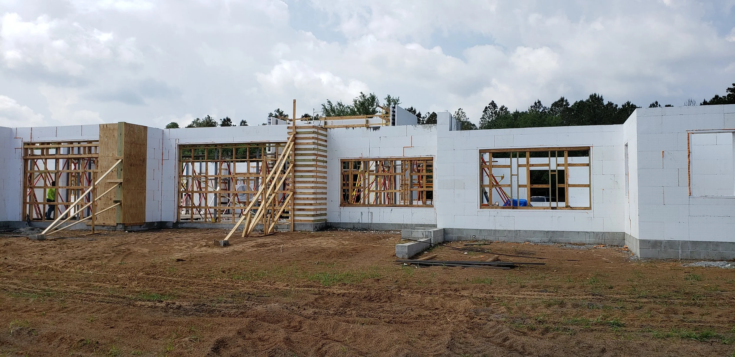 Under-construction building with wooden framing, windows, and exterior walls partially finished, on a dirt ground with cloudy sky overhead.