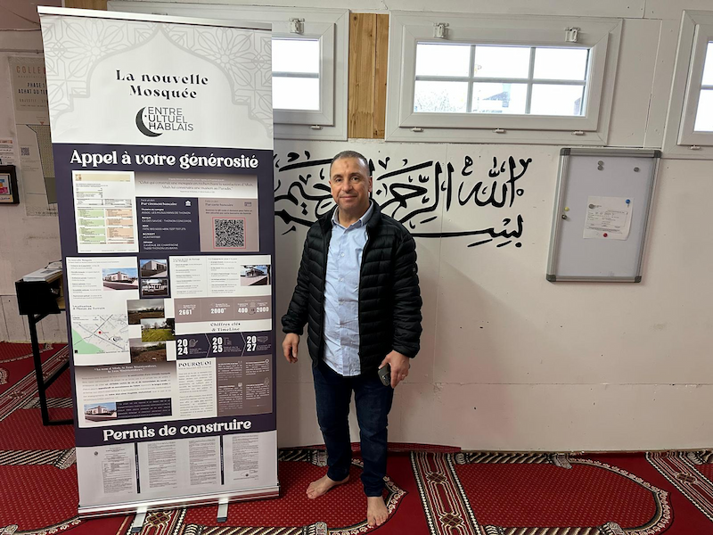 Un homme debout près d'un panneau d'affichage dans une mosquée, avec une calligraphie arabe sur le mur derrière lui.