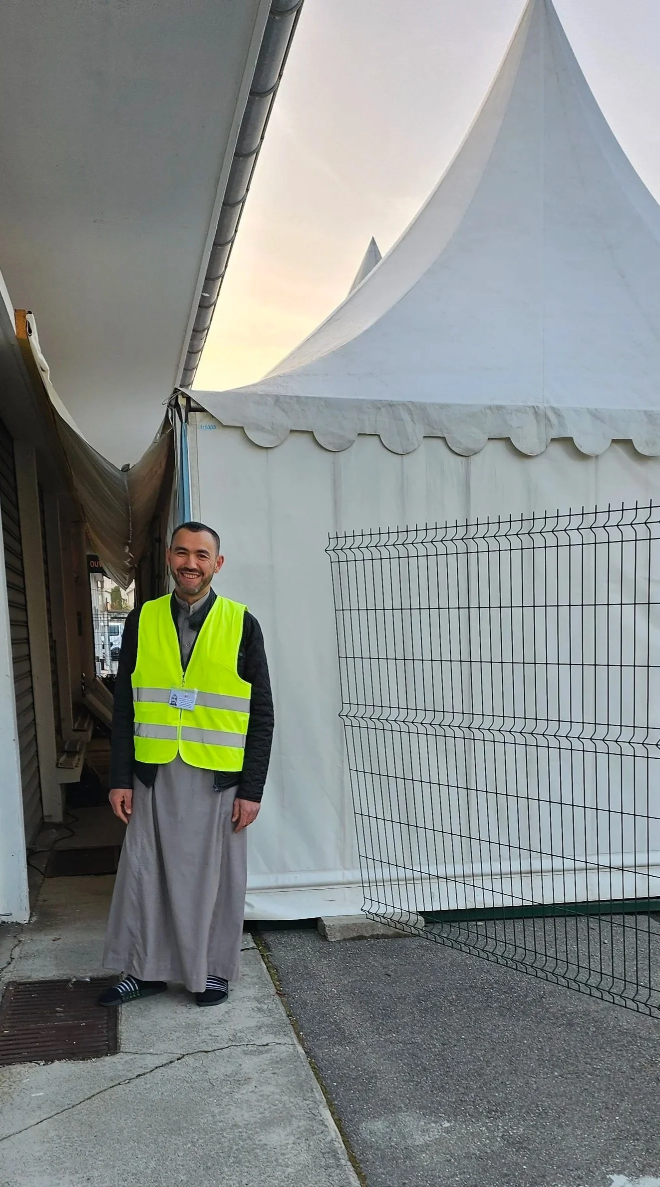 Un homme en vêtements traditionnels musulmans avec un gilet de haute visibilité, souriant, devant une tente blanche et un panneau de clôture métallique.