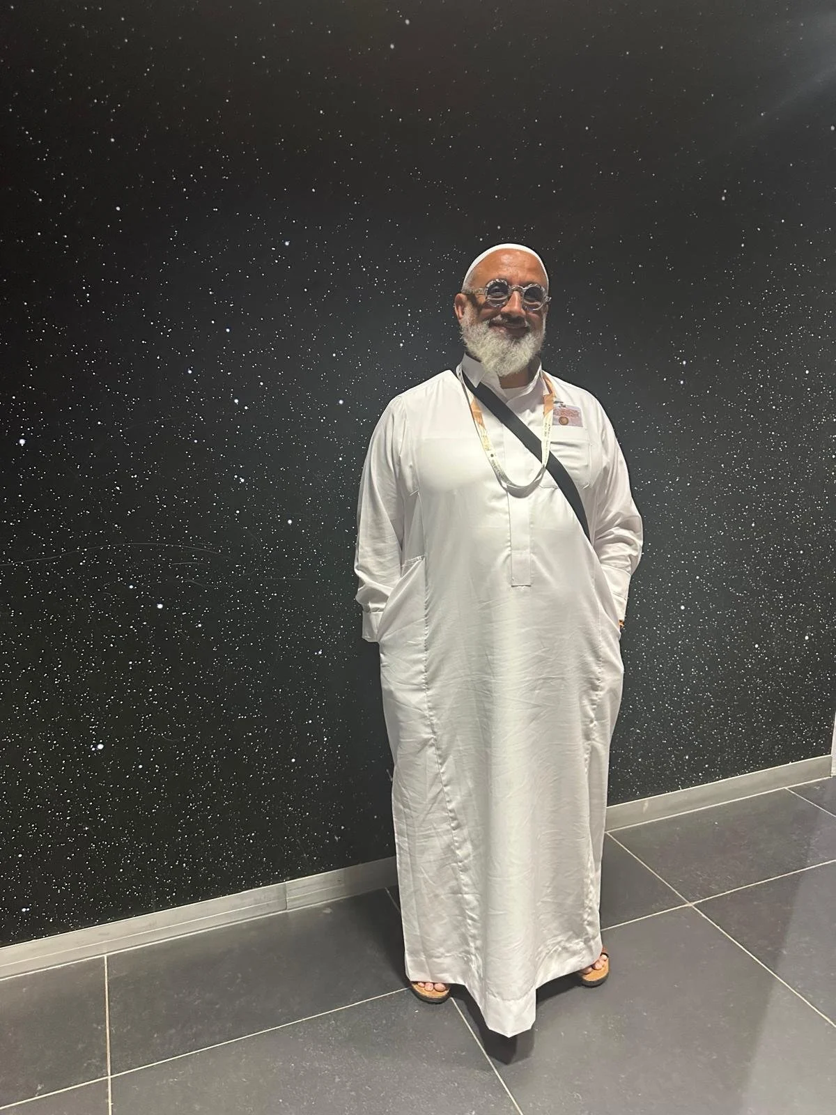 Homme souriant portant une robe traditionnelle blanche, avec des lunettes de soleil, une barbe blanche, et un badge, posé devant un mur noir avec des étoiles blanches.