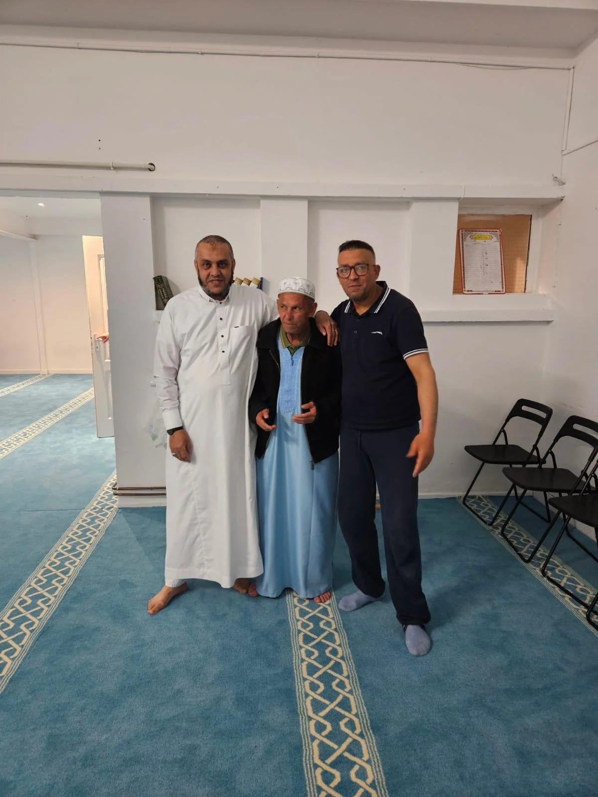 Trois hommes se tenant côte à côte dans une pièce avec un tapis bleu et des chaises noires alignées contre le mur