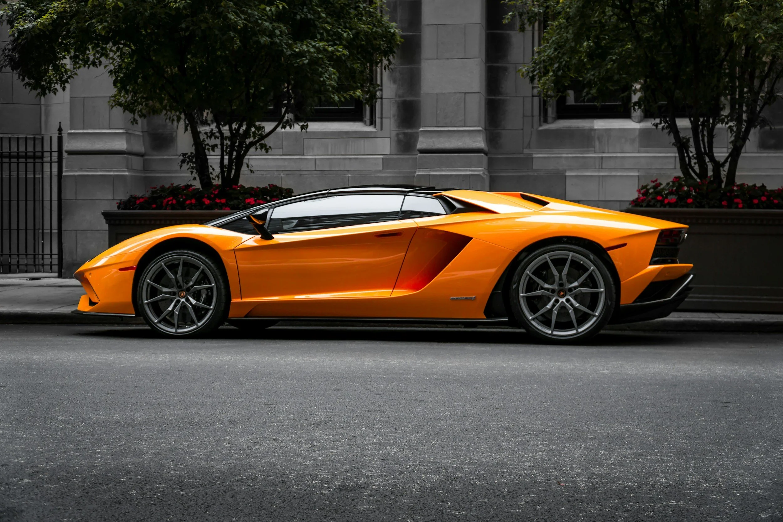 An orange Lamborghini sports car parked on an urban city street, with a gray building and trees in the background.