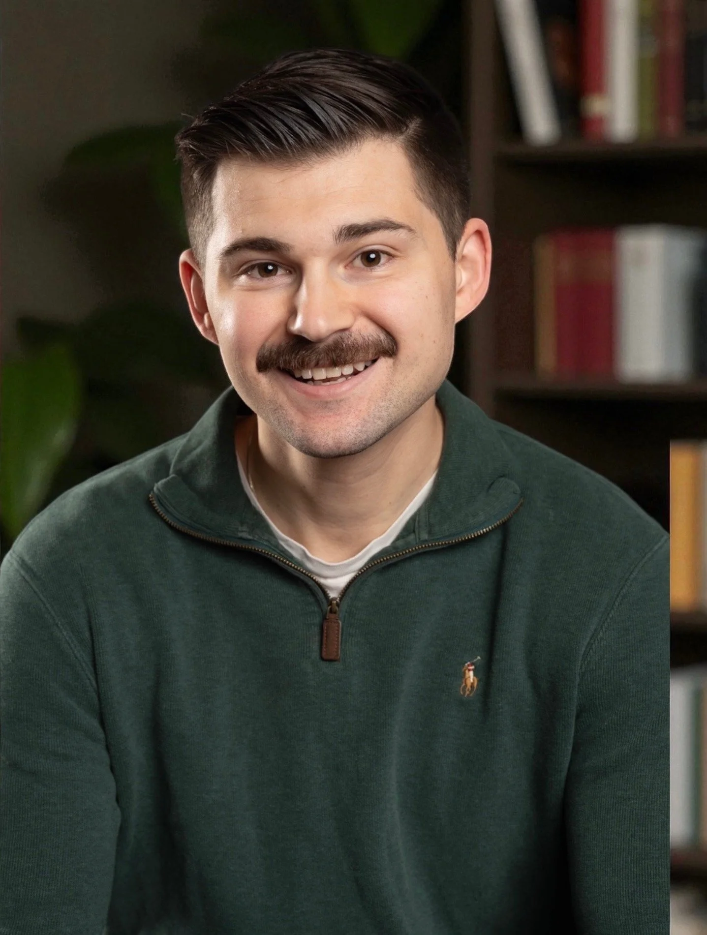 A young man with short dark hair and a mustache, smiling, wearing a dark green quarter-zip sweater, sitting indoors with a bookshelf and a plant in the background.