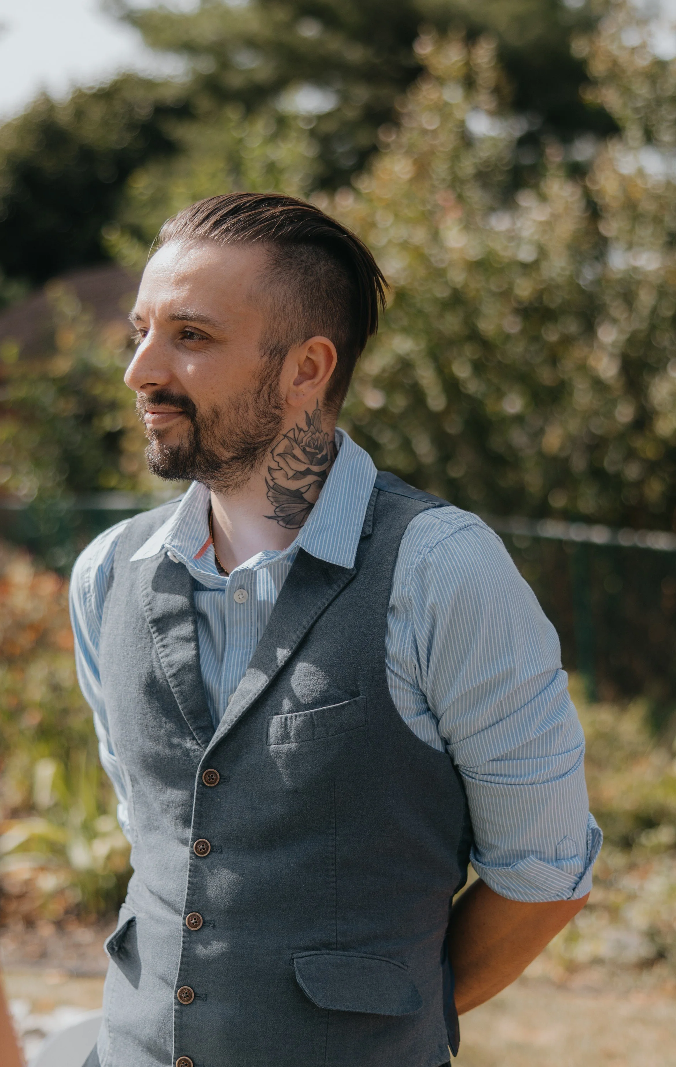A man outdoors with a tattooed neck, wearing a blue striped shirt and gray vest, looking to the side with blurred trees in the background.