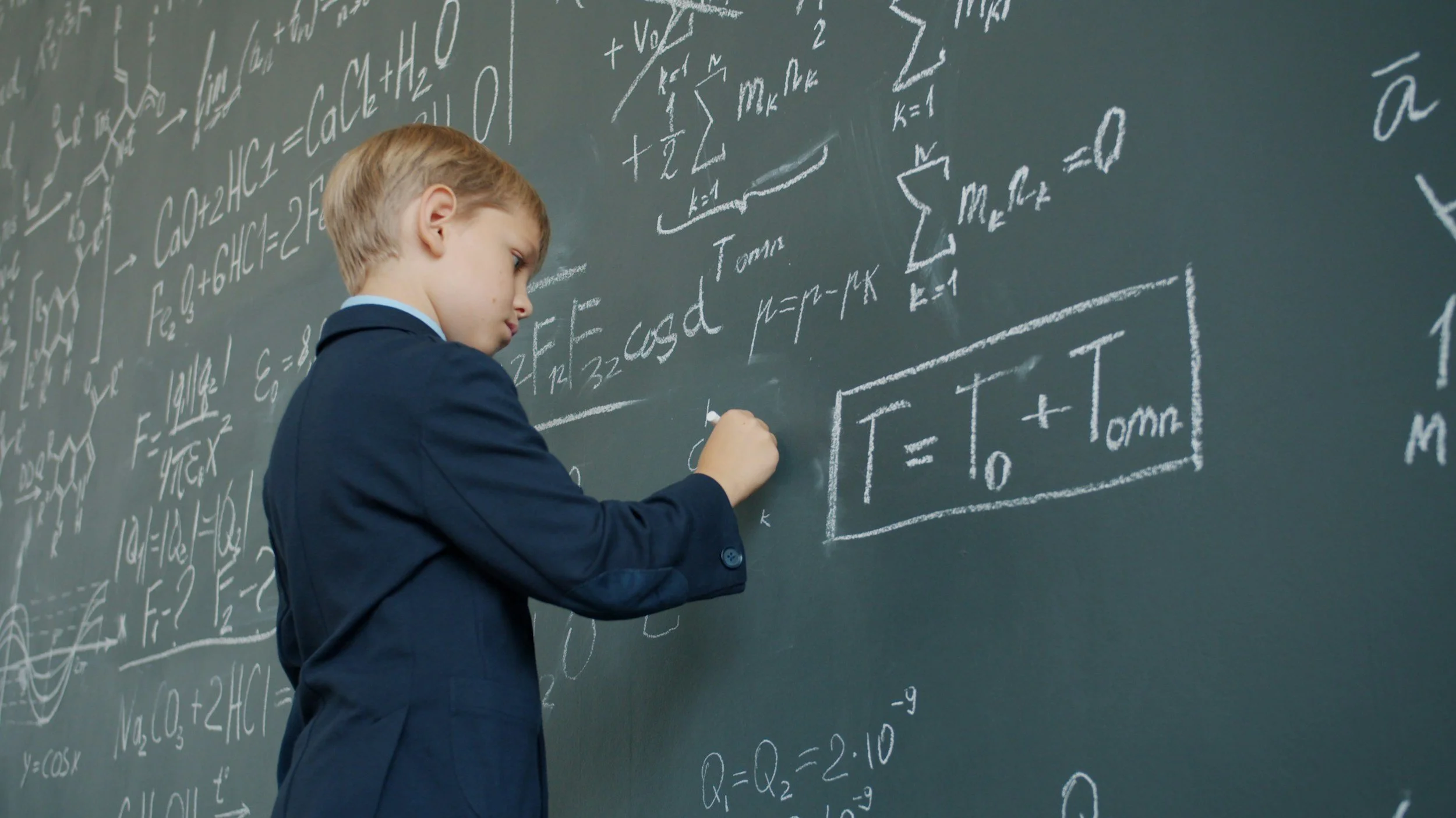 A young boy in a dark blue suit writes on a chalkboard filled with mathematical formulas and equations.