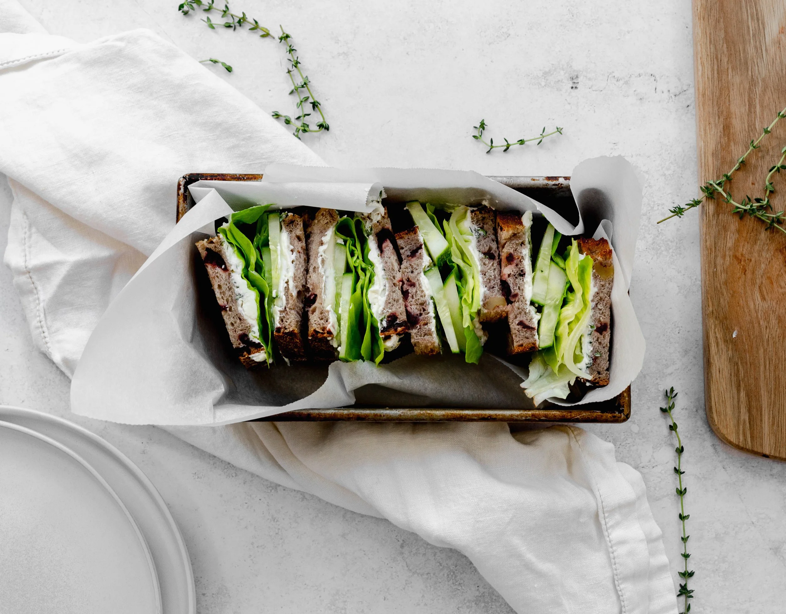 A vegetable sandwich with lettuce, cucumber, and cream cheese on whole grain bread, sliced into several pieces and placed in a rectangular baking dish lined with parchment paper, on a white surface with a wooden cutting board and greenery around.