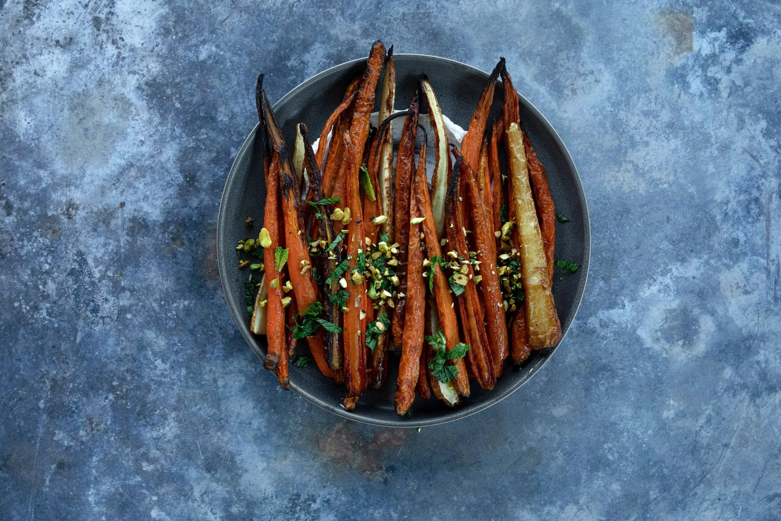 A black plate filled with roasted carrots garnished with chopped herbs and crushed nuts, on a textured gray surface.