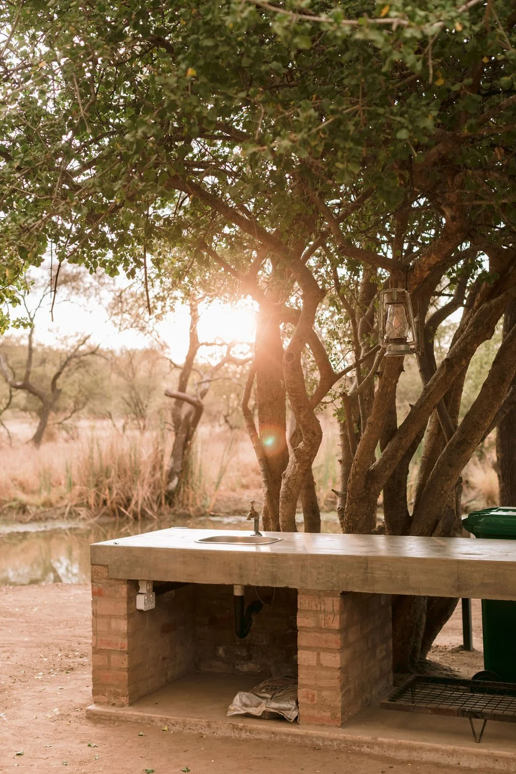 Outdoor kitchen with a brick base and wooden countertop next to trees with green foliage. The sun is setting, shining through the branches, with a lantern hanging from a tree branch. A small sink is built into the counter, and there are some items un