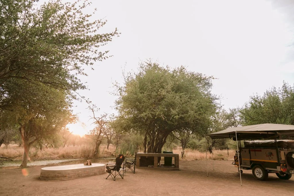 Camping scene in a natural setting with trees, a small pond, camping chairs, a picnic table, and a golf cart with a canopy, during sunset or early evening.