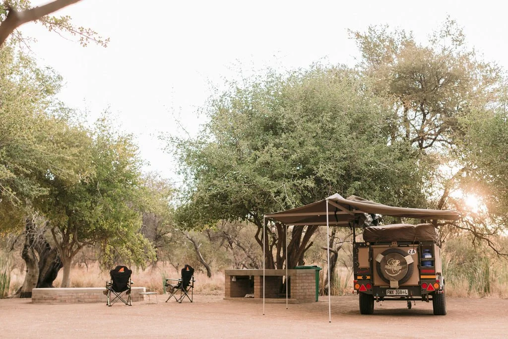 Outdoor camping area with a vehicle, two chairs, and a canopy next to trees at sunset or sunrise.