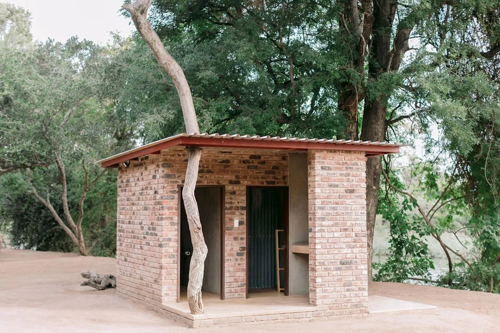 Small brick structure with a corrugated metal roof, situated outdoors among trees, with a white, twisted tree trunk growing through the building.