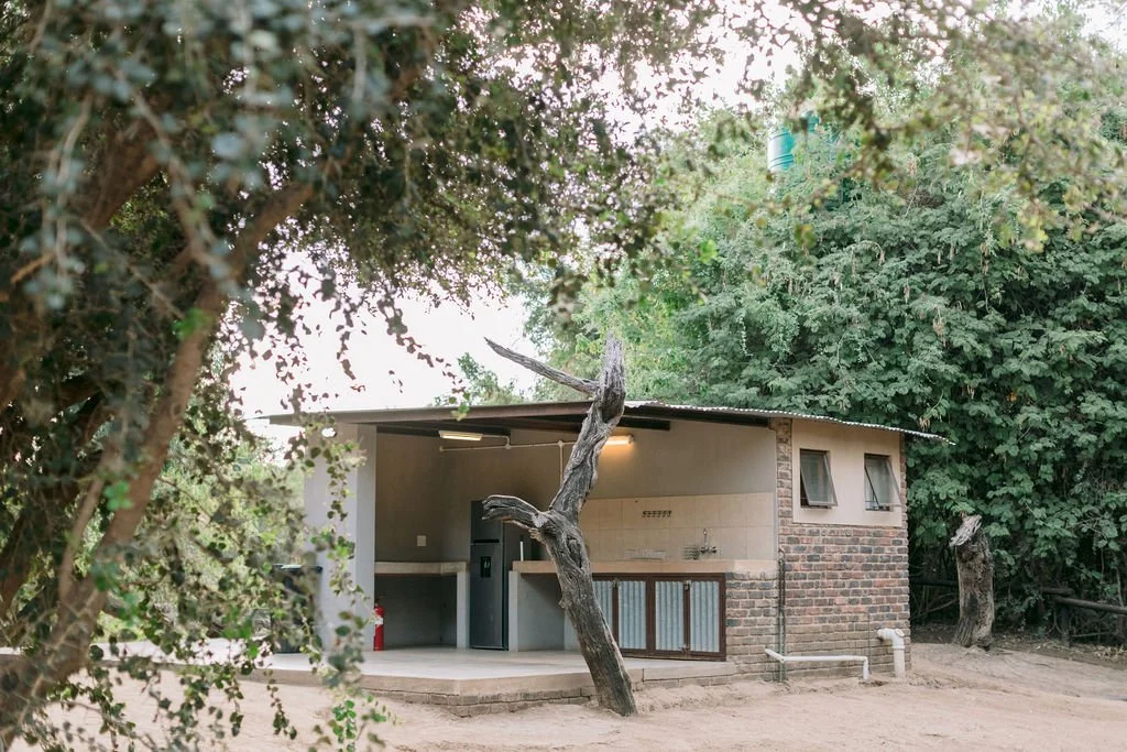 Small building with brick and concrete walls, surrounded by dense green trees and bushes, with an outdoor kitchen area.