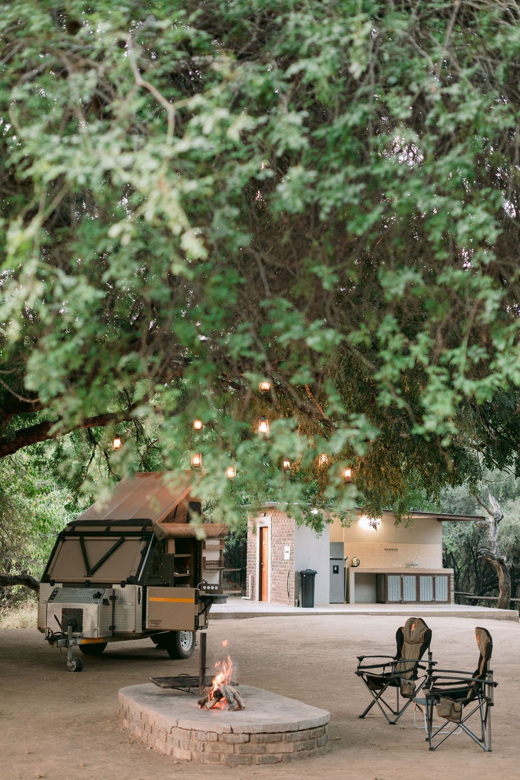 Outdoor camping scene showing a large tree with string lights, a fire pit with a small fire, two folding chairs, a mobile food trailer, and a small building in the background.
