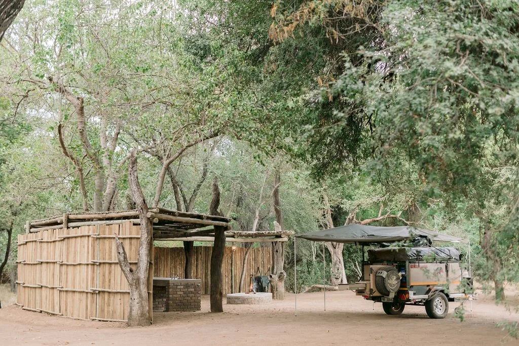 A rustic outdoor scene features a wooden seating area with a roof, trees around, and an off-road trailer with a canopy parked on dirt ground.