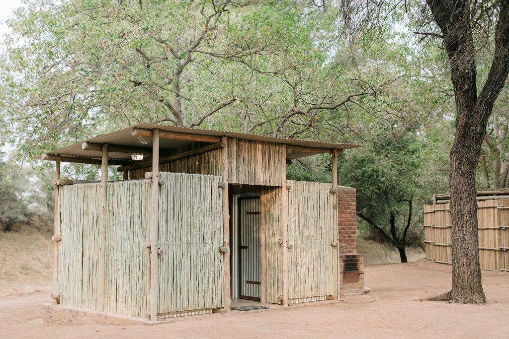 A rustic outdoor shower area with bamboo walls and a metal roof, situated on dirt ground with trees and a wooden fence in the background.