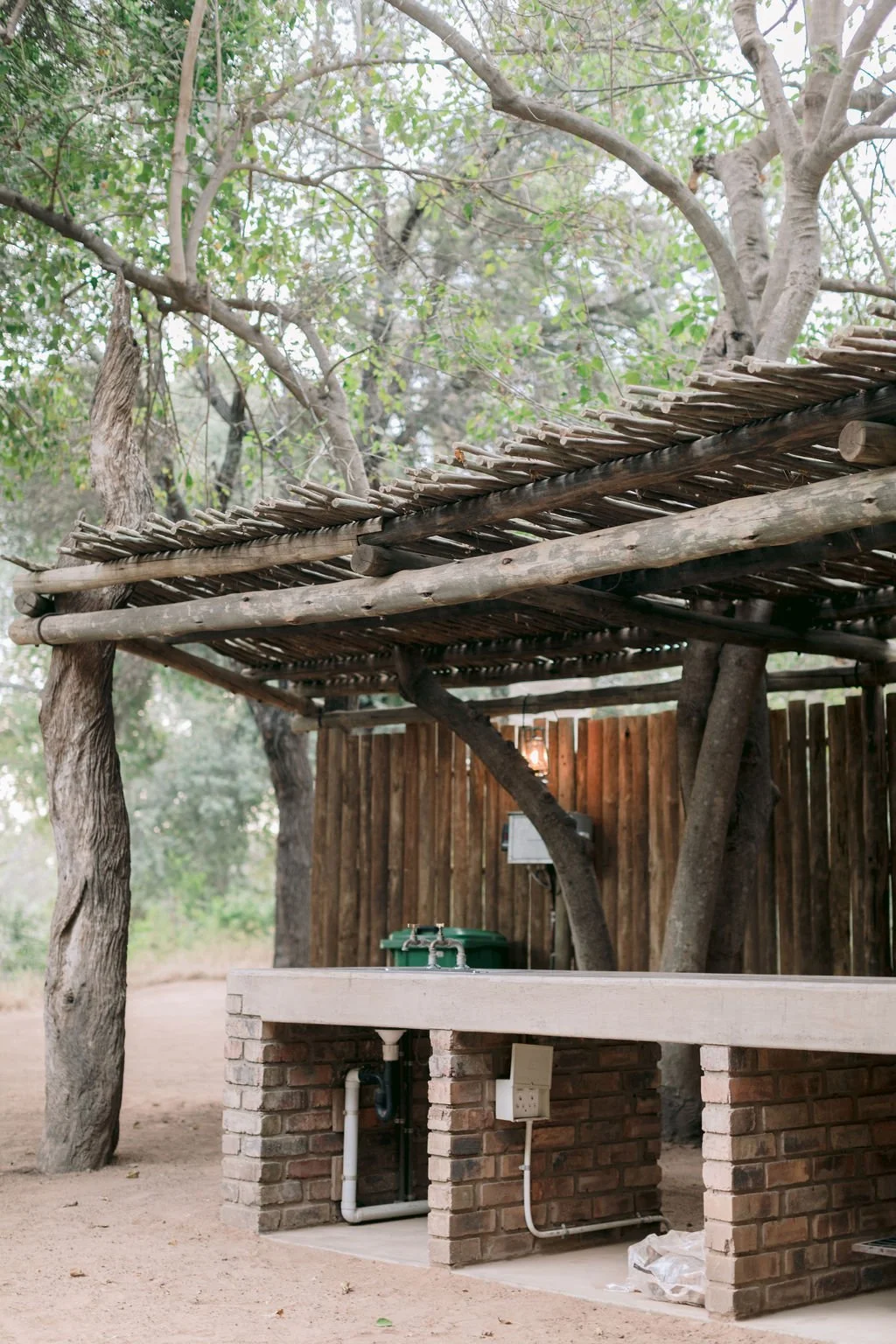 Outdoor rustic shelter with a wooden roof supported by tree branches and trunks, featuring a brick countertop with a green container, electrical outlets, and exposed pipes, surrounded by trees.